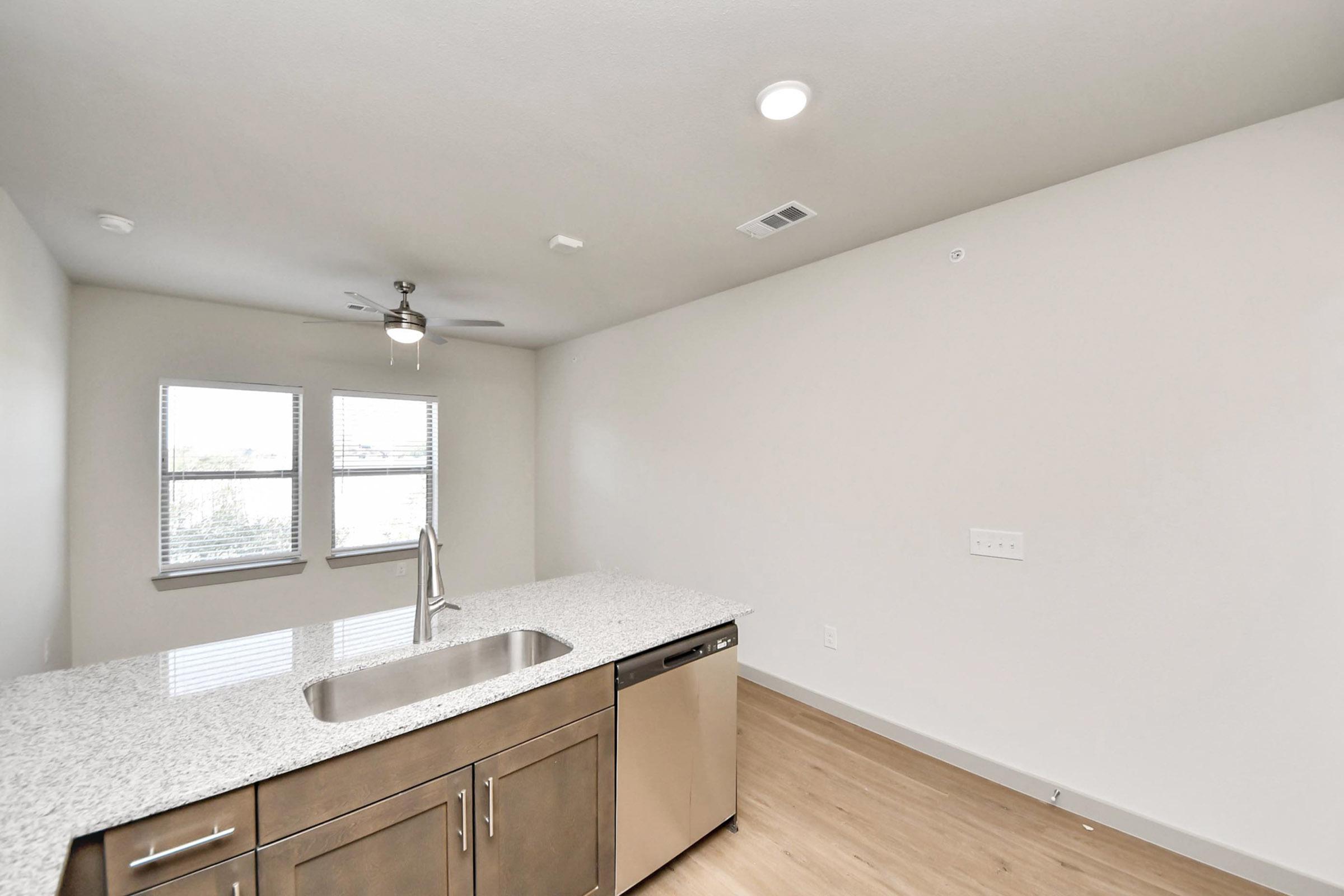 Modern kitchen interior featuring a granite countertop island with a sink and stainless steel dishwasher. Large windows allow natural light to fill the space, complemented by a ceiling fan. The walls are painted a light color, enhancing the bright and airy atmosphere of the room. Wood-look flooring completes the design.