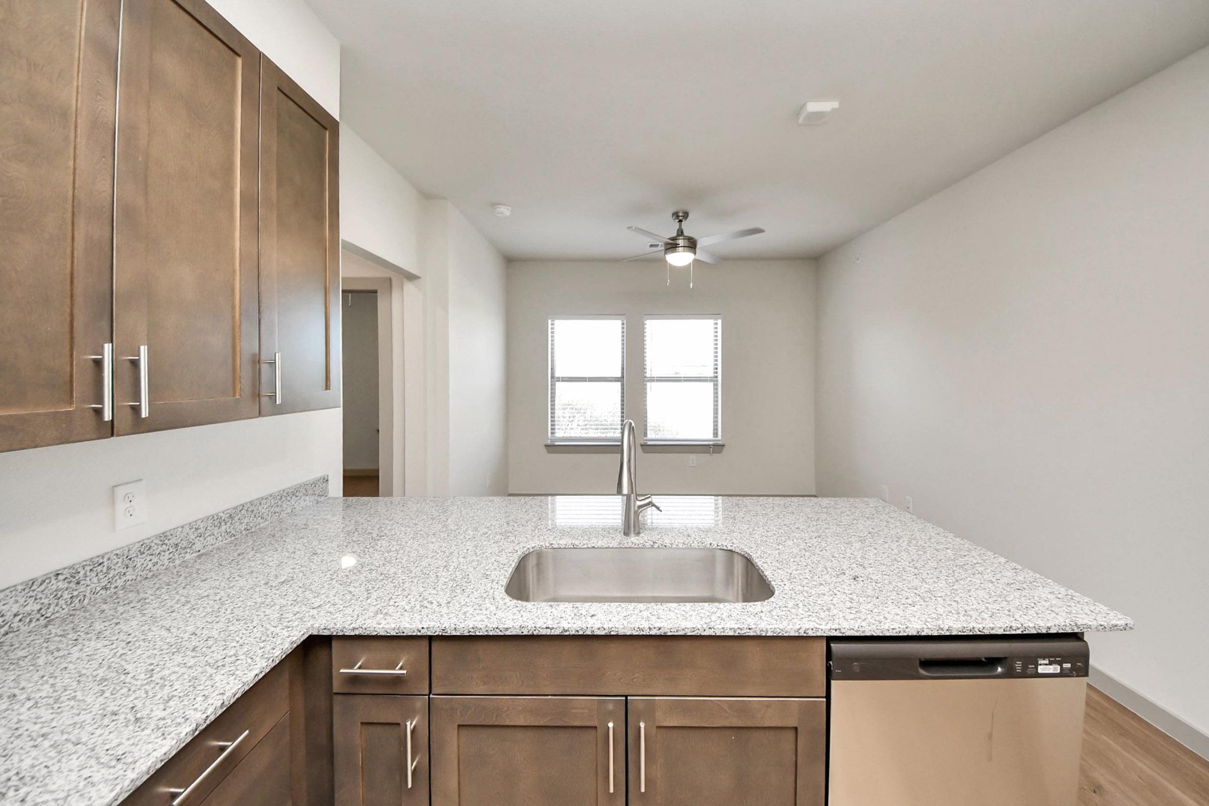 Modern kitchen view featuring a granite countertop with a stainless steel sink, a dishwasher, and wooden cabinets. Bright and spacious with a ceiling fan and large windows allowing natural light, connecting to a living area in the background. Neutral wall colors enhance the airy atmosphere.