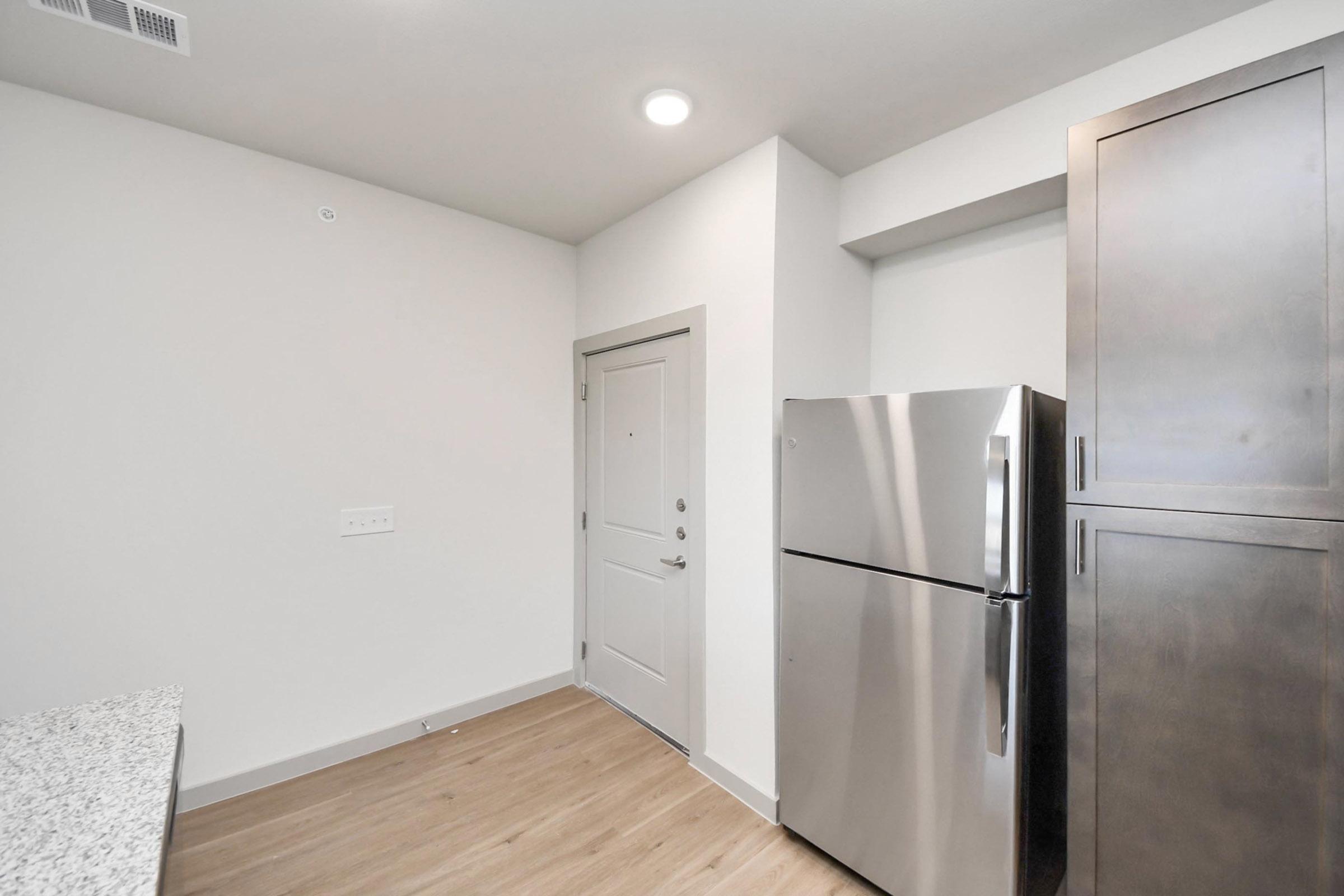 Modern kitchen area featuring a stainless steel refrigerator, beige walls, and light wood flooring. The corner displays a front door with a lock and light fixtures above. Cabinets in dark wood are visible beside the fridge, complementing the contemporary design.