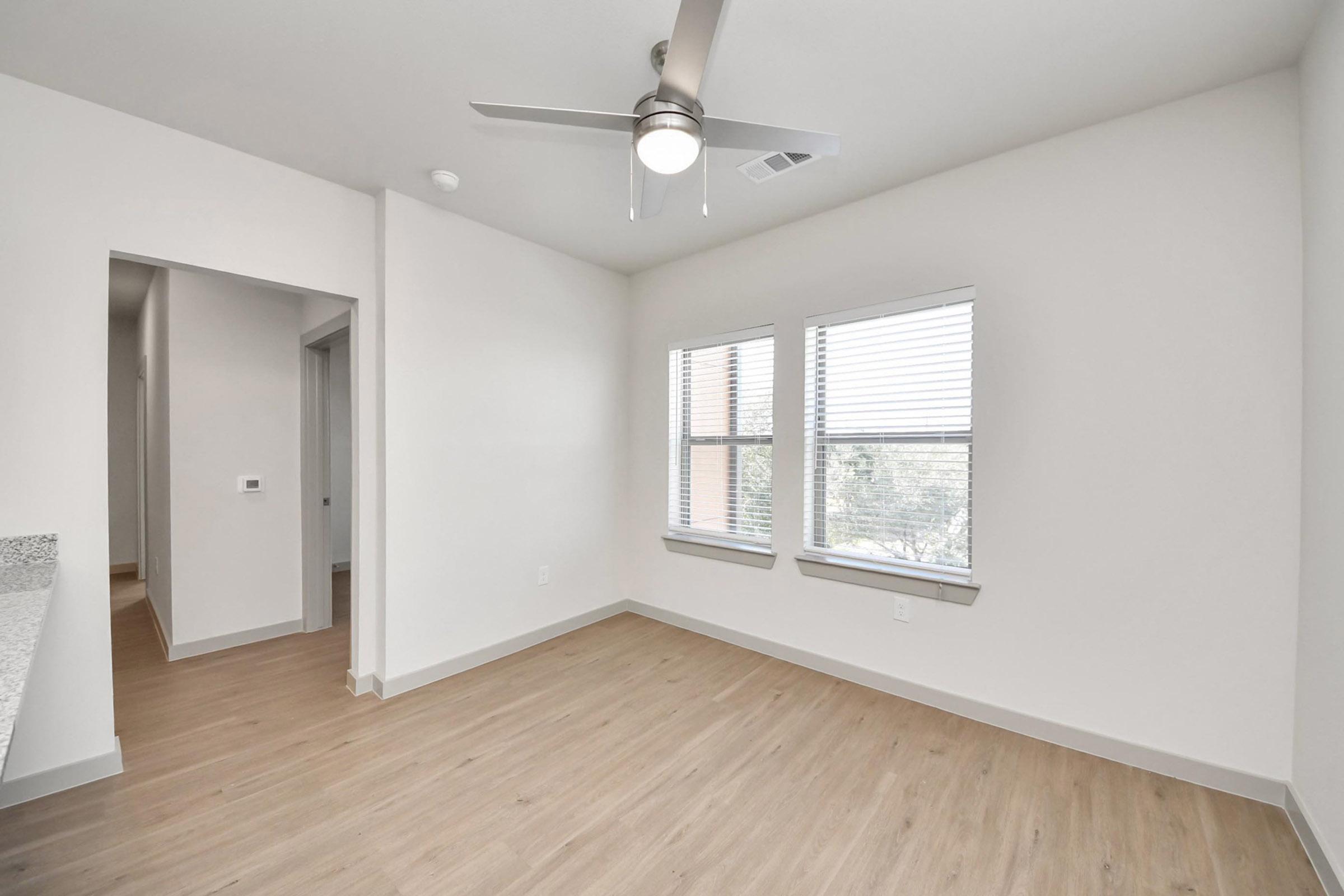 A bright, modern room featuring light wood flooring, a ceiling fan, and two windows with blinds. One wall has an open doorway leading to another area, and there are clean, neutral-colored walls. The room is designed with a contemporary aesthetic and offers natural light.