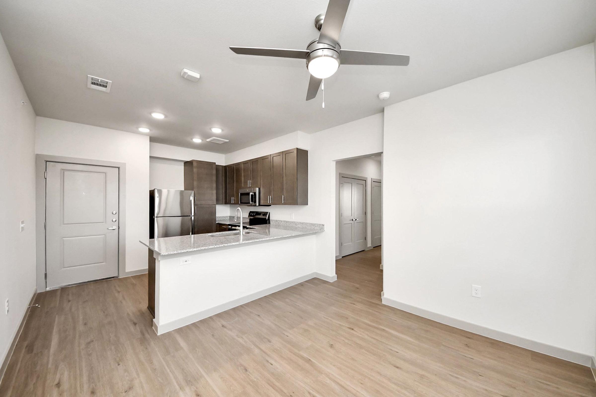 Modern kitchen and living area in an apartment. Features include light-colored walls, hardwood floors, stainless steel appliances, a ceiling fan, and granite countertops. The space is open, with a doorway leading to another room visible in the background.