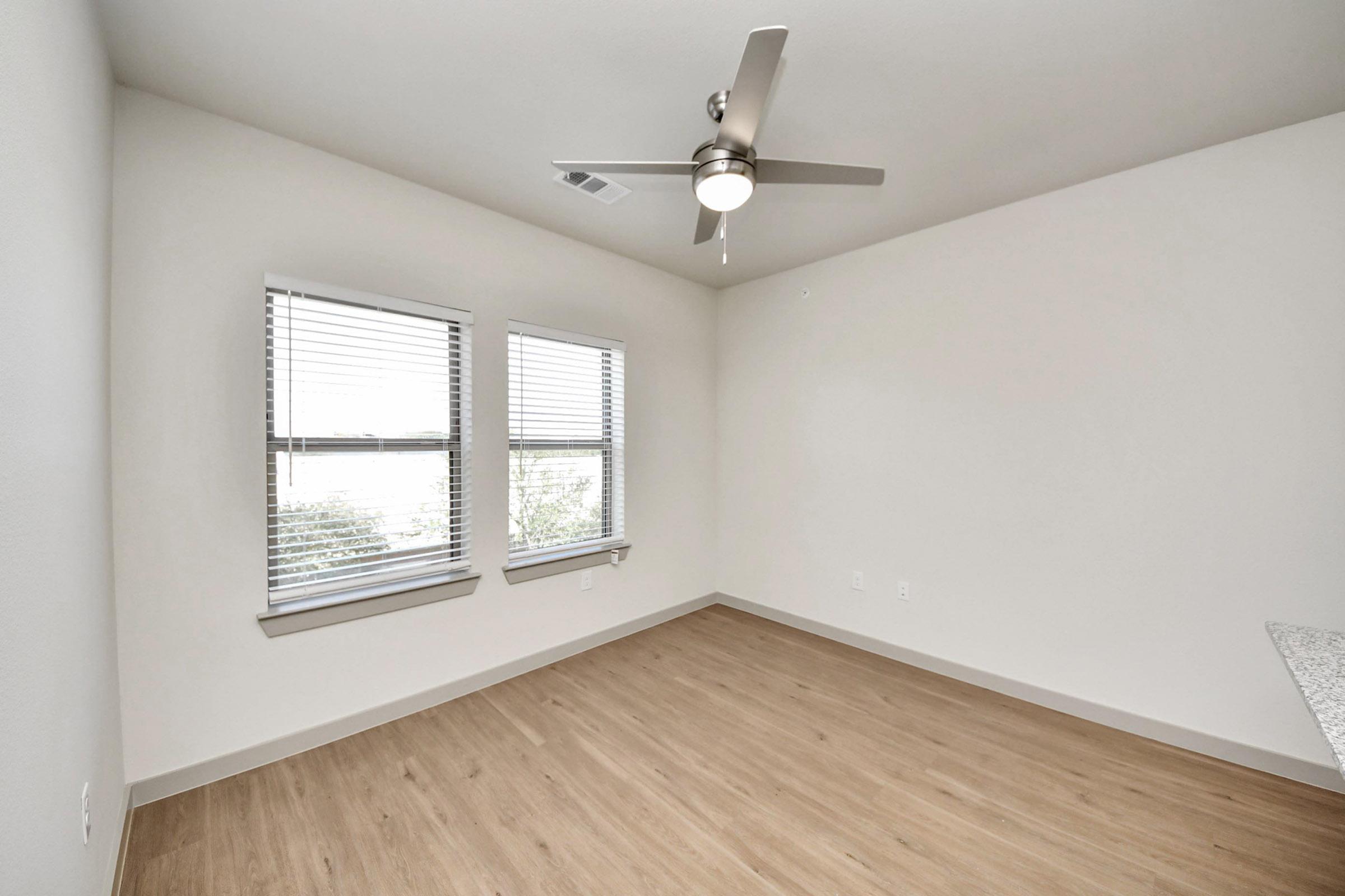 A minimalistic empty room featuring light-colored walls, two windows with blinds, and a ceiling fan. The flooring is light wood laminate, and there’s a small countertop visible in one corner. The space is bright and airy, giving a sense of openness.