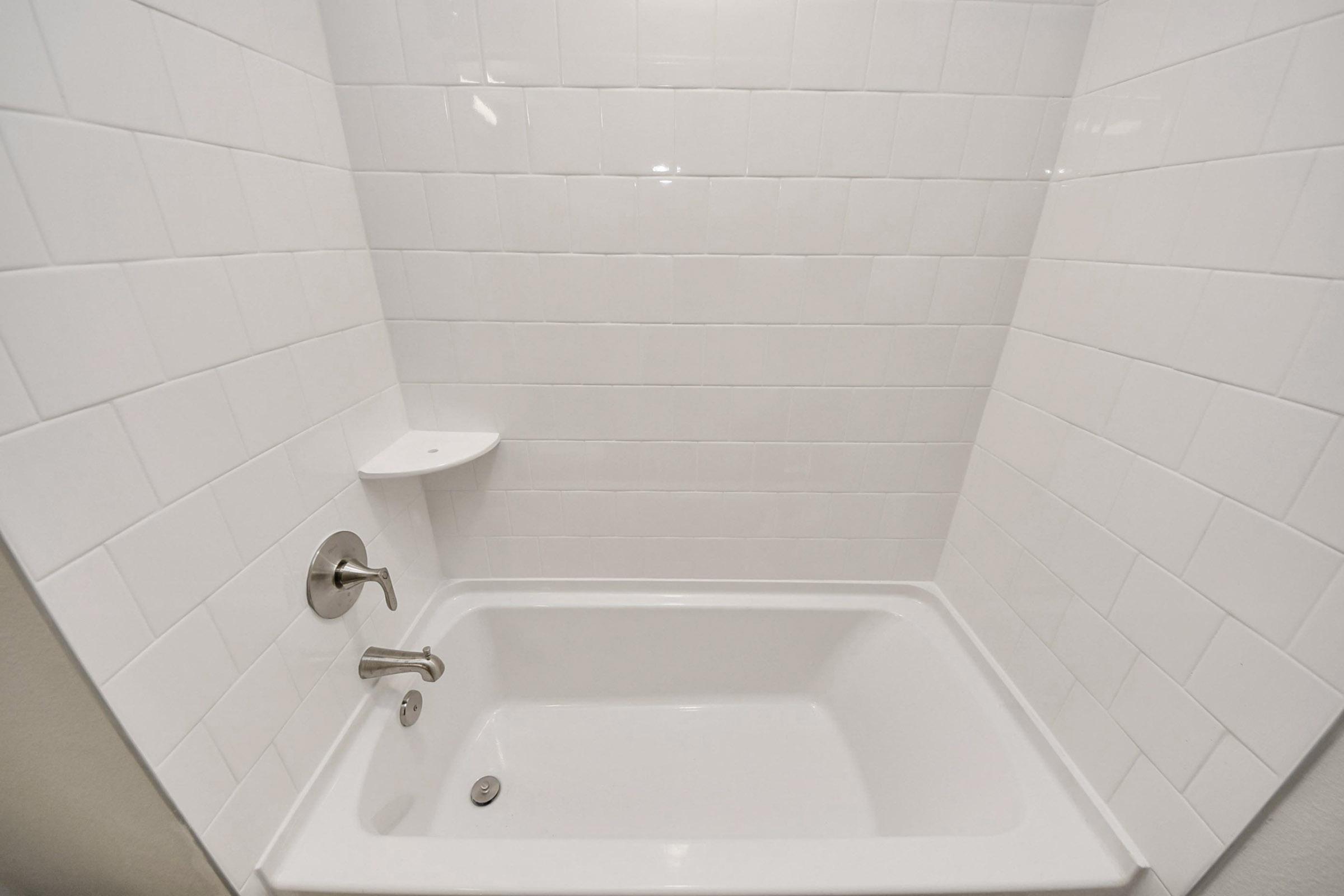 A clean, modern bathtub with a white tiled surround. The bathtub features a chrome faucet and a separate showerhead attachment. There is a small shelf built into the wall for toiletries. The overall color scheme is bright and minimalist, creating a spacious feel.