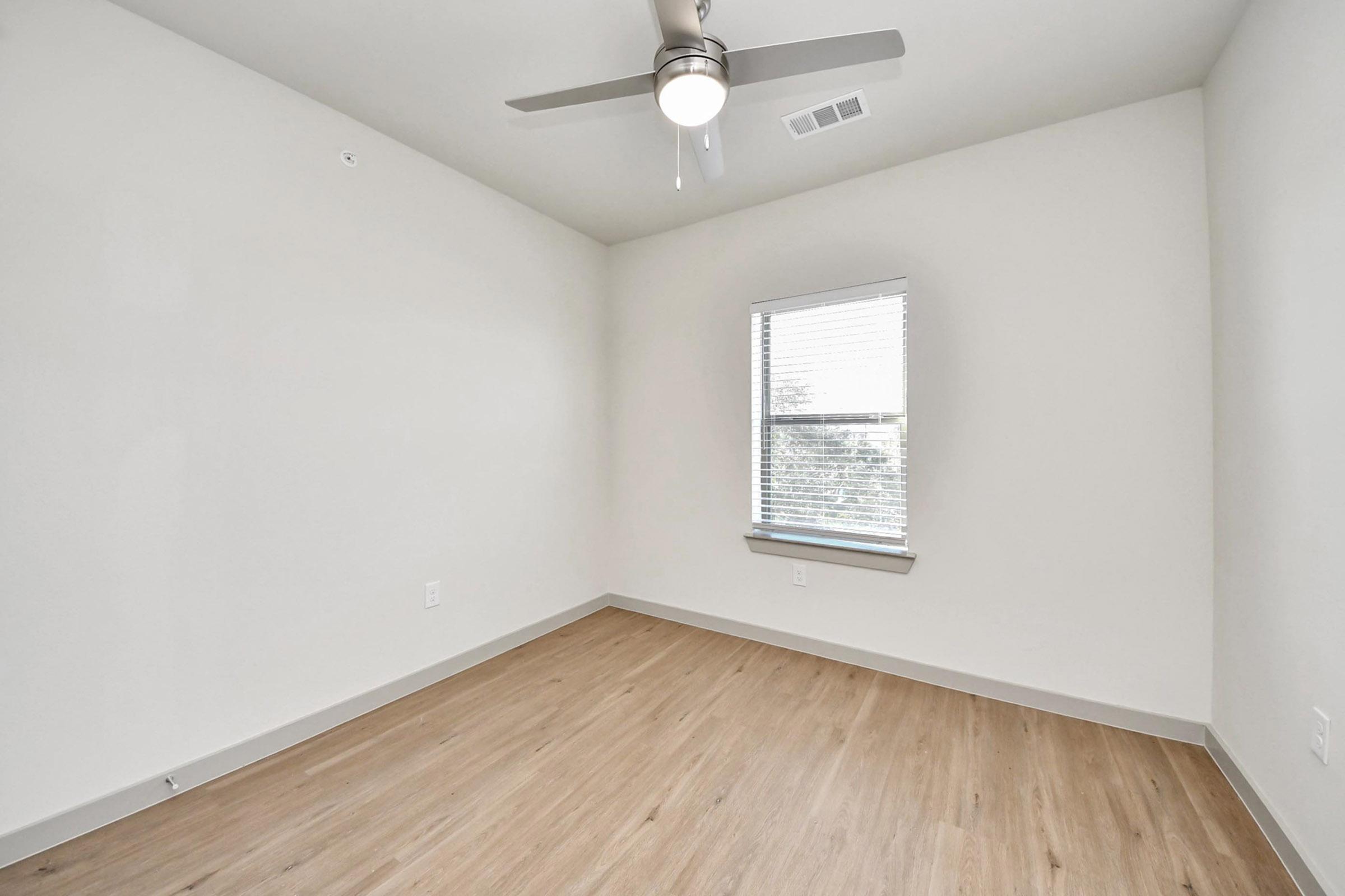A sparsely furnished room with light-colored walls and wood laminate flooring. It features a ceiling fan and a single window with blinds, allowing natural light to enter. The room has a clean, minimalist look with ample space for furniture.