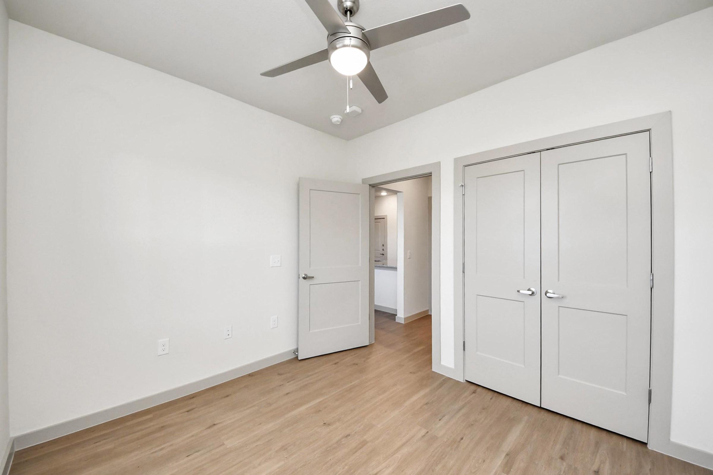 A modern, empty room featuring light wood flooring and white walls. Two closed doors are visible, one leading to a closet and the other to a hallway. A ceiling fan is mounted above, providing a contemporary touch to the space. Natural light comes from the open doorway, giving the room an airy feel.