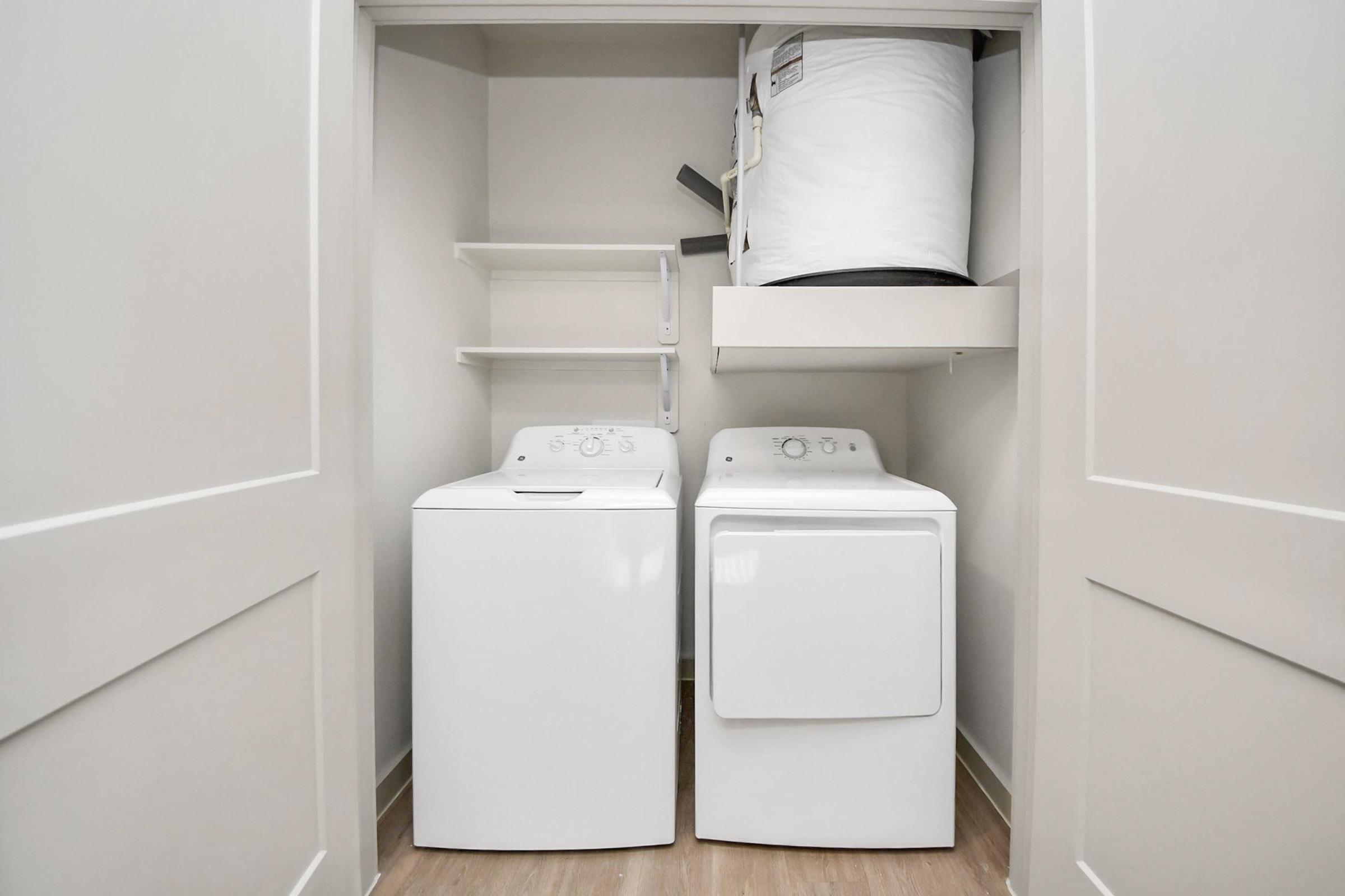 A small laundry area featuring a stacked washer and dryer, along with shelves for storage. The appliances are white, and the room has light-colored walls and wooden flooring, creating a clean and functional space for laundry tasks.