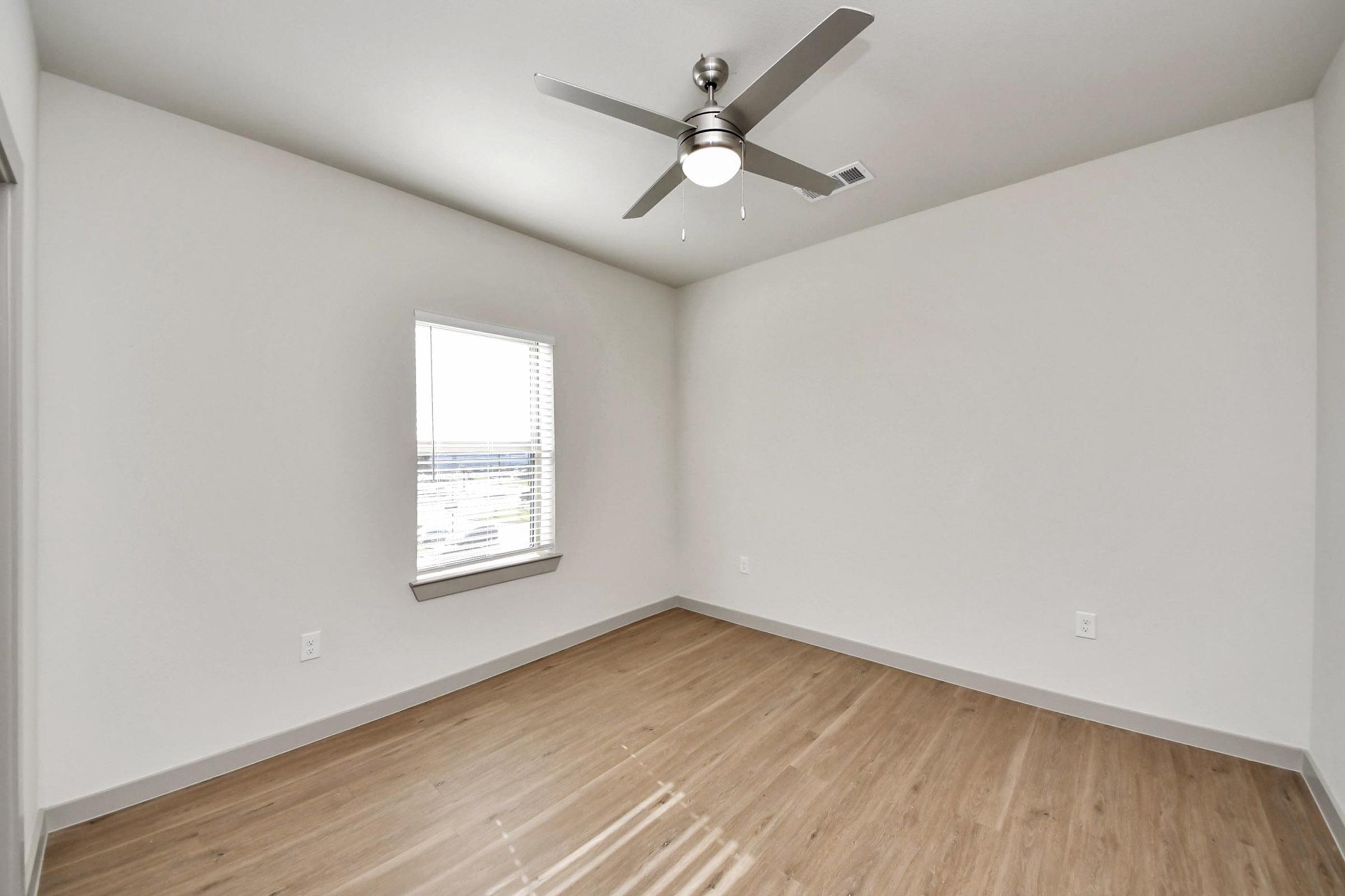 A minimalistic, empty room with light-colored walls and wooden flooring. A single window allows natural light to enter, and a ceiling fan is mounted in the center. The space is tidy and lacks furniture, emphasizing its spaciousness.