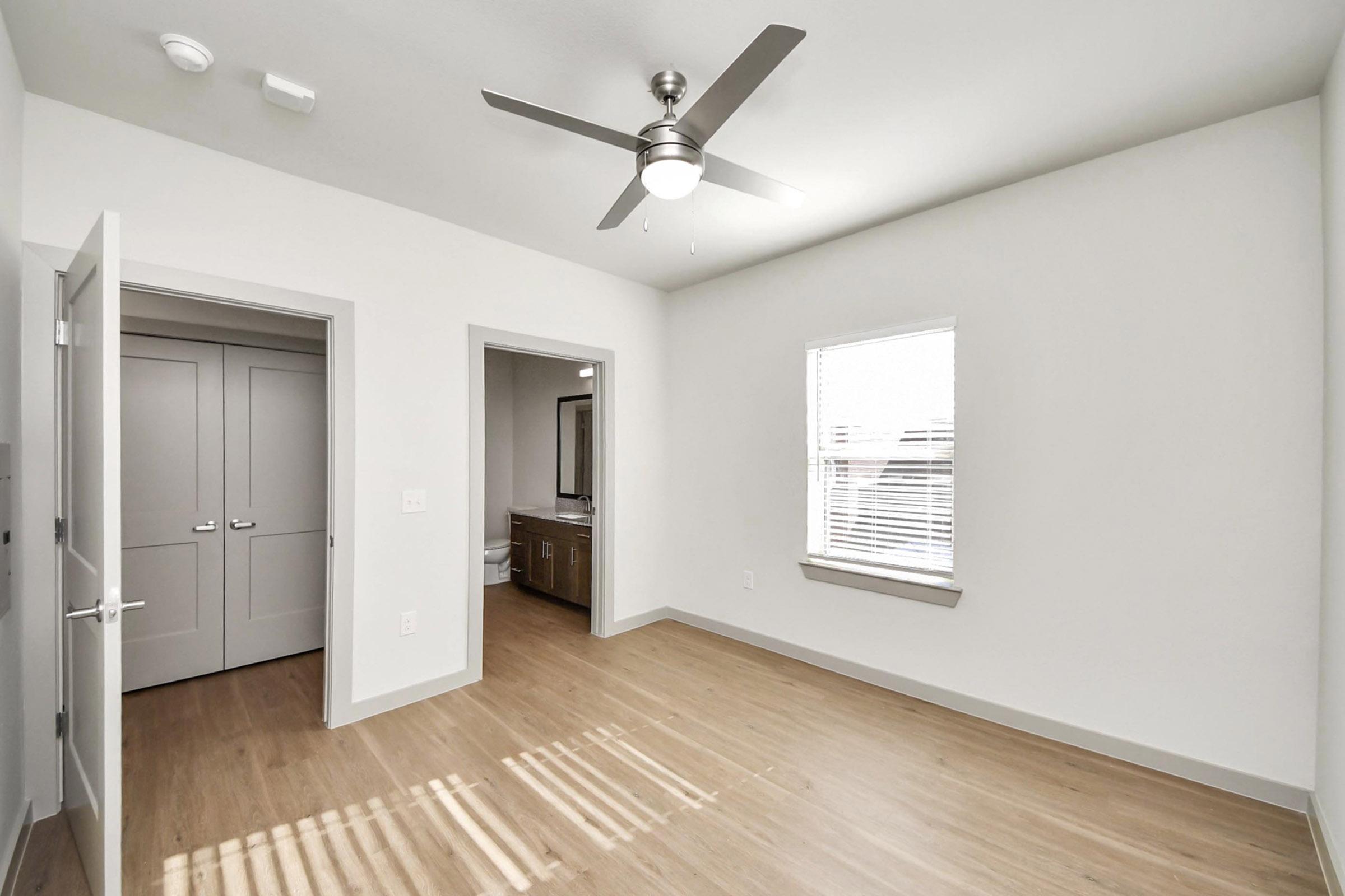 An empty room featuring light-colored walls and wooden flooring. A ceiling fan is installed, and a window brings in natural light. To the left, there is an open door leading to another room, and a closet door can be seen straight ahead. The space is bright and airy, suggesting a modern interior design.