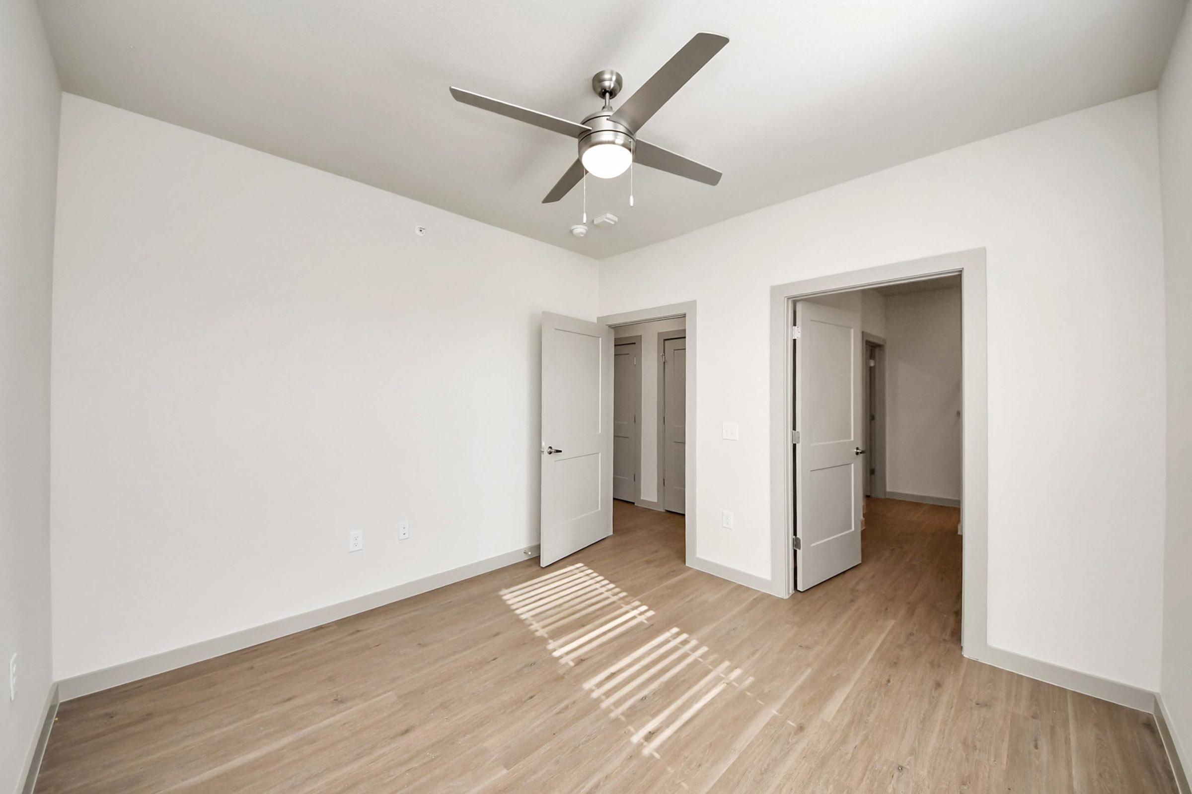 A bright, empty room with light-colored walls and wood-like flooring. It features a ceiling fan and two doorways leading to other spaces. Natural light casts shadows on the floor from a nearby window, adding warmth to the minimalist aesthetic.