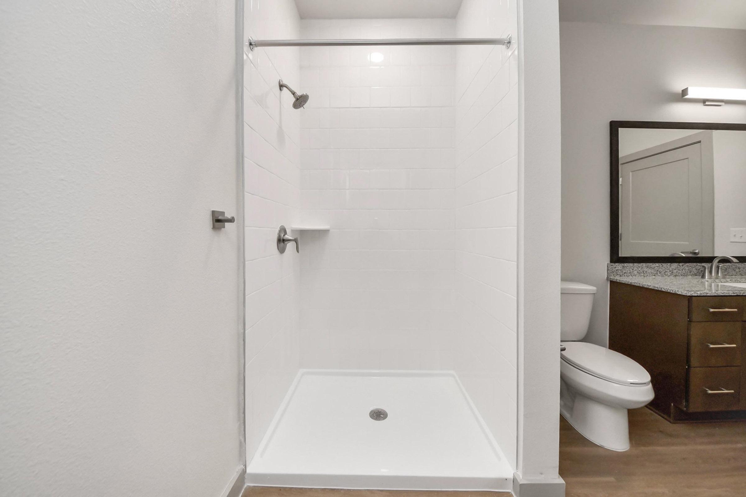 A clean, modern bathroom featuring a walk-in shower with a glass door, white tiled walls, and a single shelf. Adjacent to the shower is a compact toilet and a vanity with a dark wood finish and a granite countertop, illuminated by a light fixture above. The flooring is light wood.