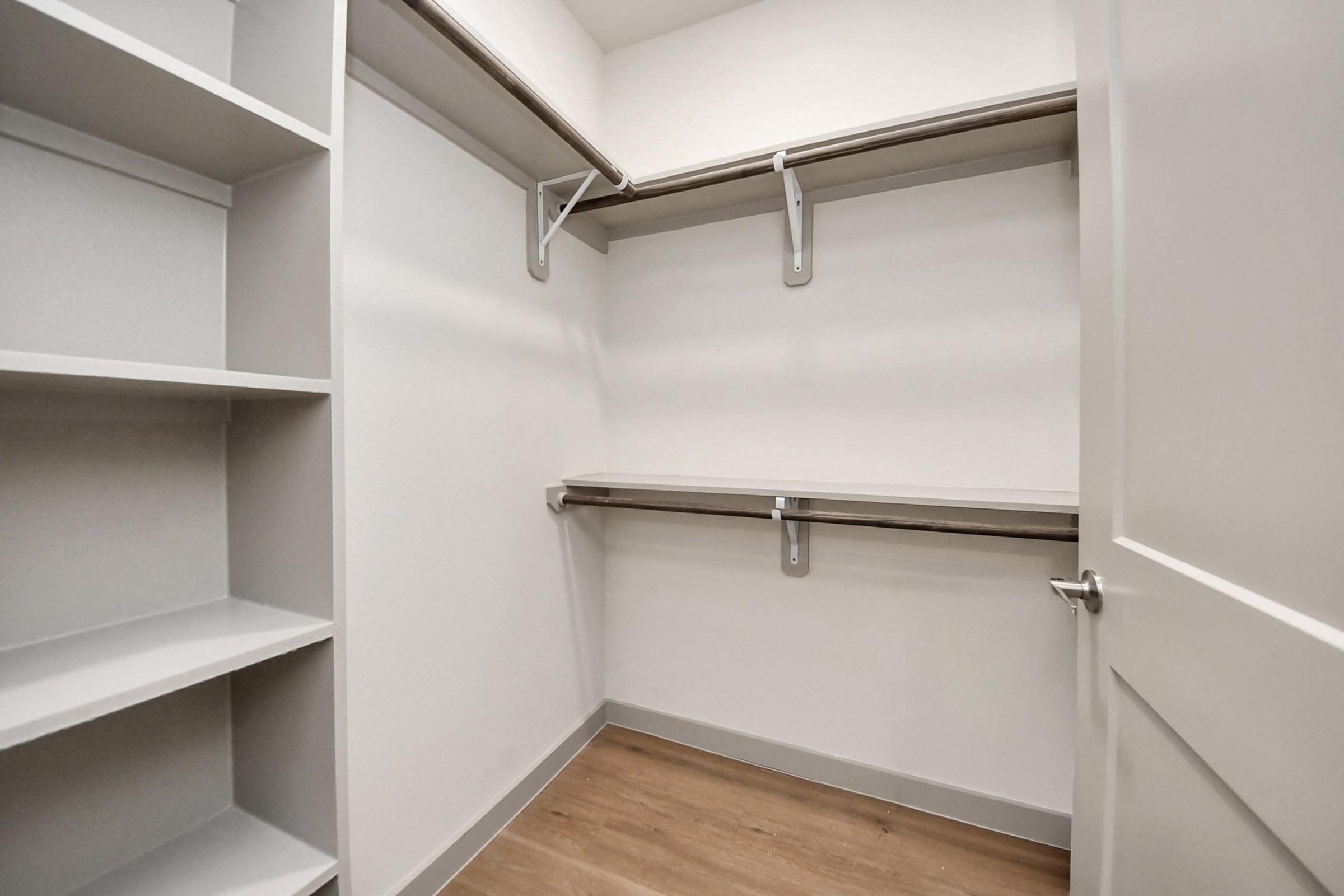 A spacious, empty closet featuring two hanging rods and a central shelf, with light-colored walls and wooden flooring. One side has built-in shelves for additional storage. The overall design is modern and minimalistic, providing ample space for organization.