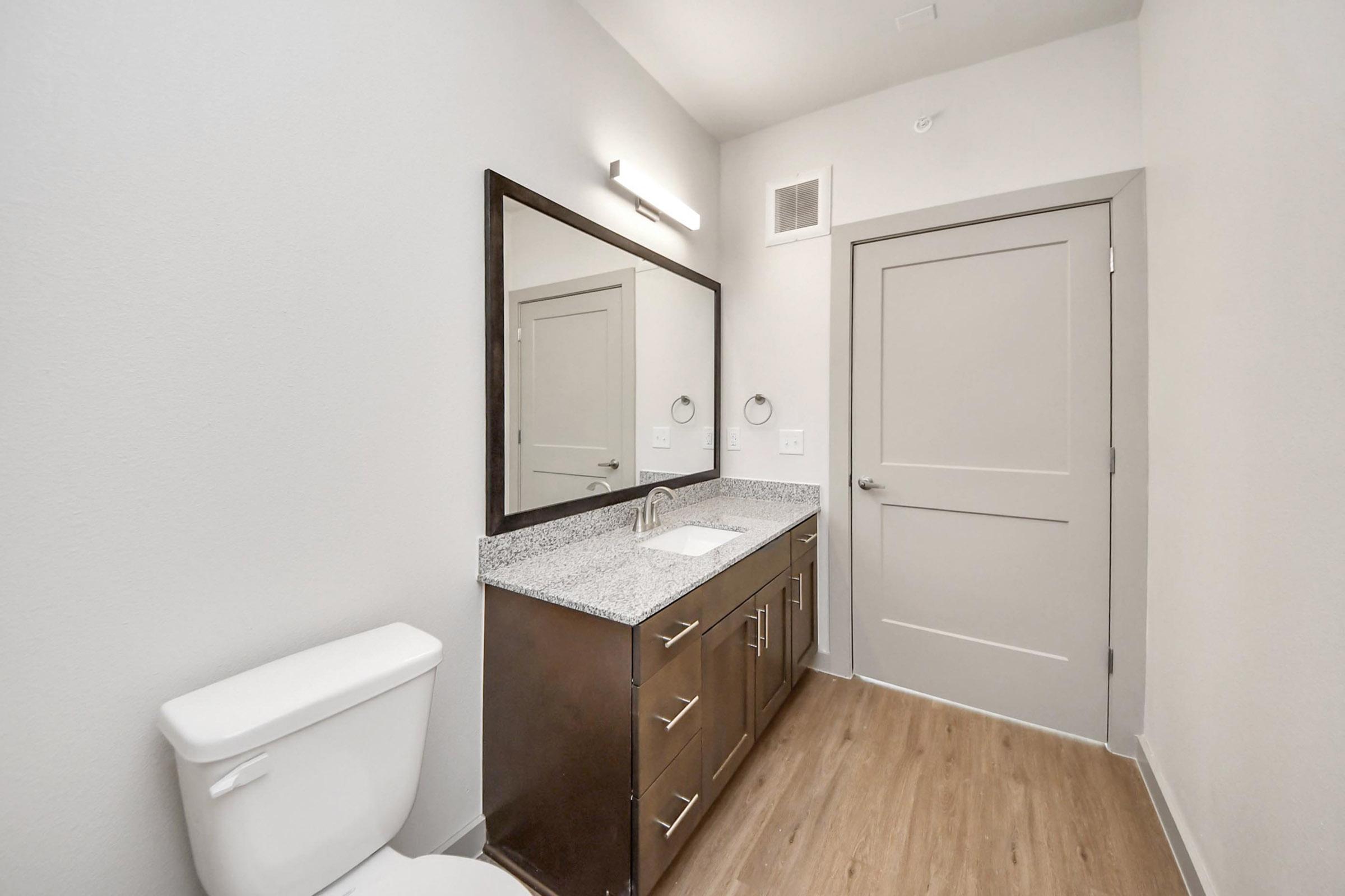 A modern bathroom featuring a white toilet, a dark wood vanity with a gray speckled countertop, and a rectangular mirror above the sink. The walls are painted light gray, and there is a door leading out of the room. The flooring is a light wood finish.