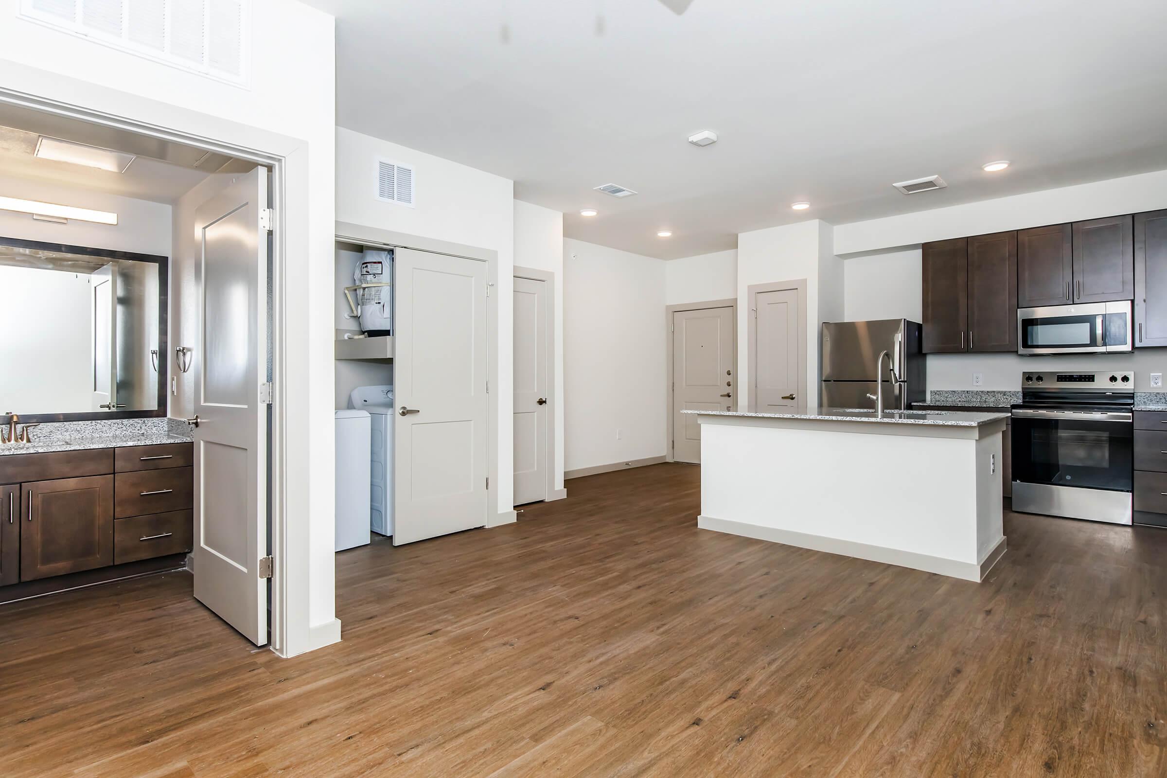 Modern kitchen and living area in a bright, open-concept space. Features hardwood-style flooring, stainless steel appliances, and dark wood cabinetry. Includes a laundry nook with a washer behind closed doors. Natural light enters through large windows, creating a welcoming atmosphere.