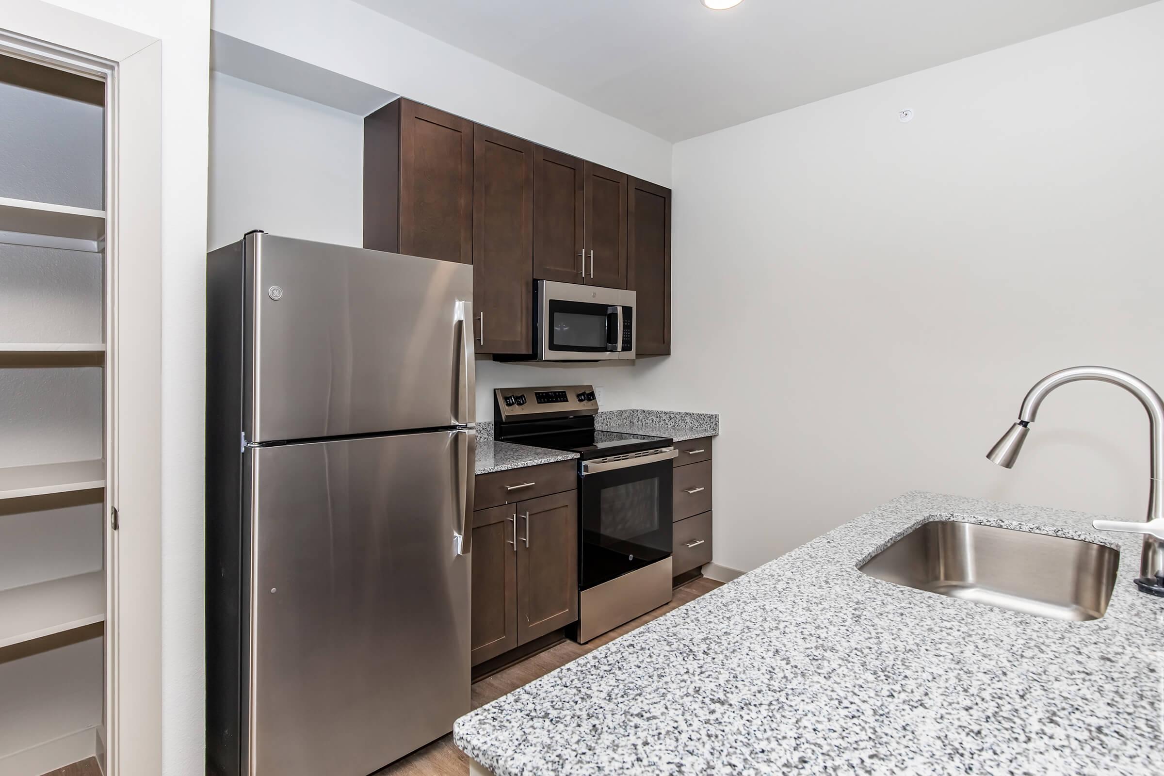 Modern kitchen featuring dark wood cabinets, stainless steel refrigerator, microwave, and oven. The countertops are speckled granite, and there's a sink with a modern faucet. A pantry cabinet is visible next to the refrigerator, with ample storage space. Bright lighting enhances the open layout.