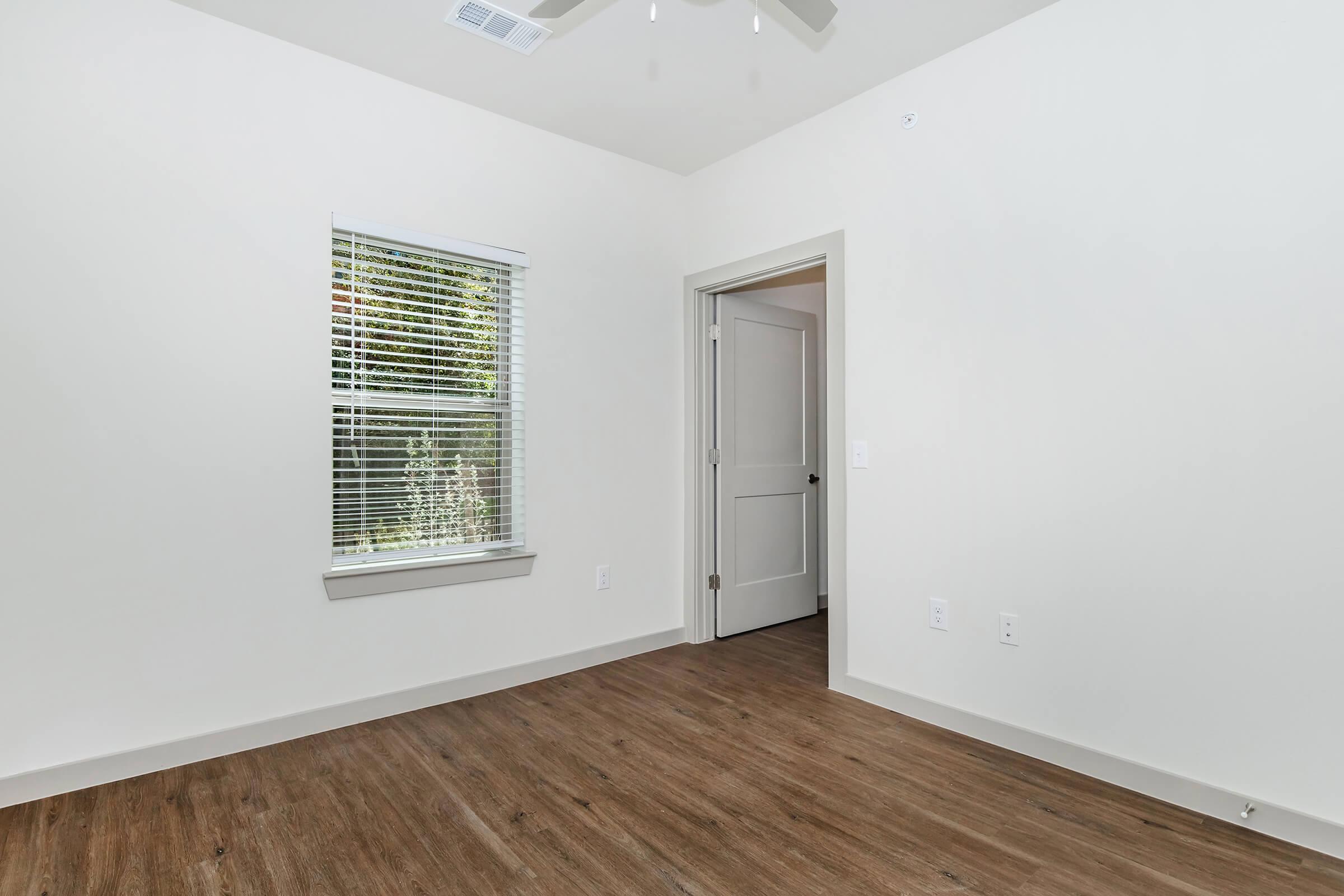 A bright, minimalist room with light-colored walls and wooden flooring. It features a window with blinds, allowing natural light to enter, and an open door leading to another space. The decor is simple and modern, creating a clean and spacious atmosphere.