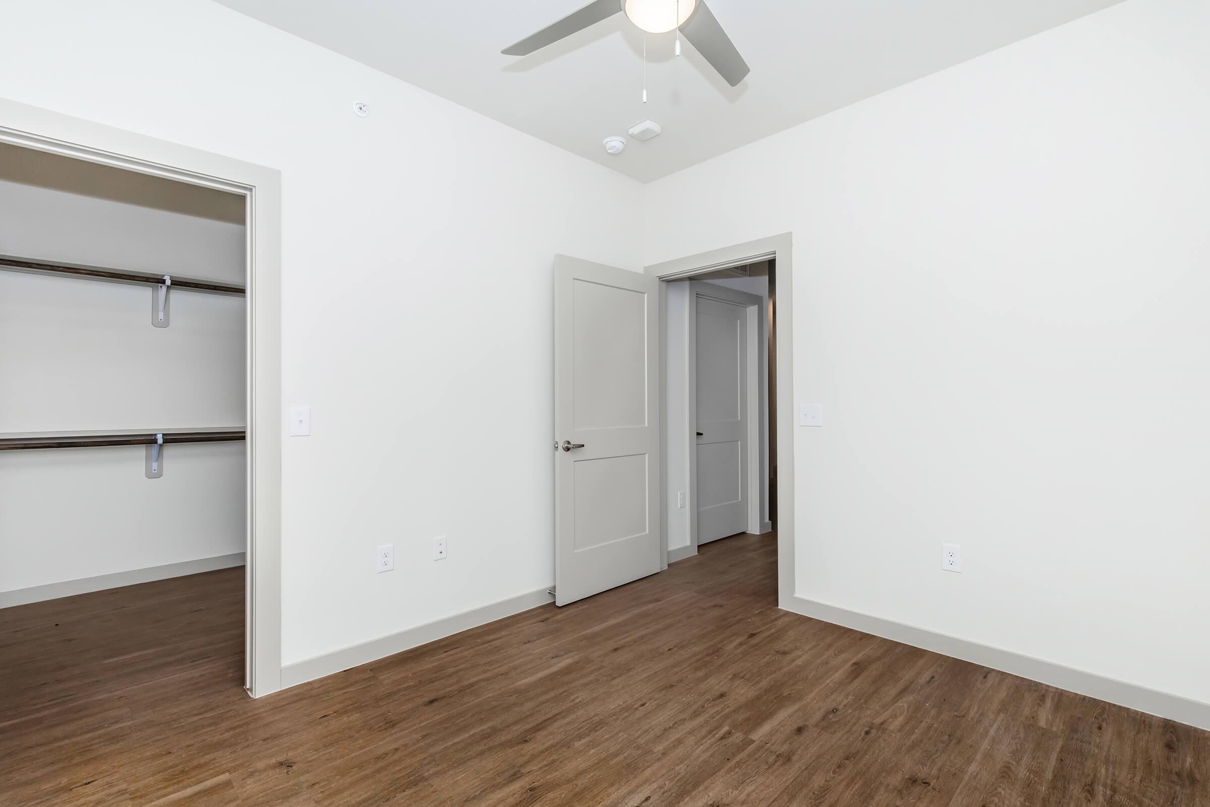 Interior of a modern room featuring light-colored walls and a ceiling fan. One side has a doorway leading to a closet with shelves. The opposite side shows a second open doorway leading to another room, with a clean, minimalist design and wood-style flooring.