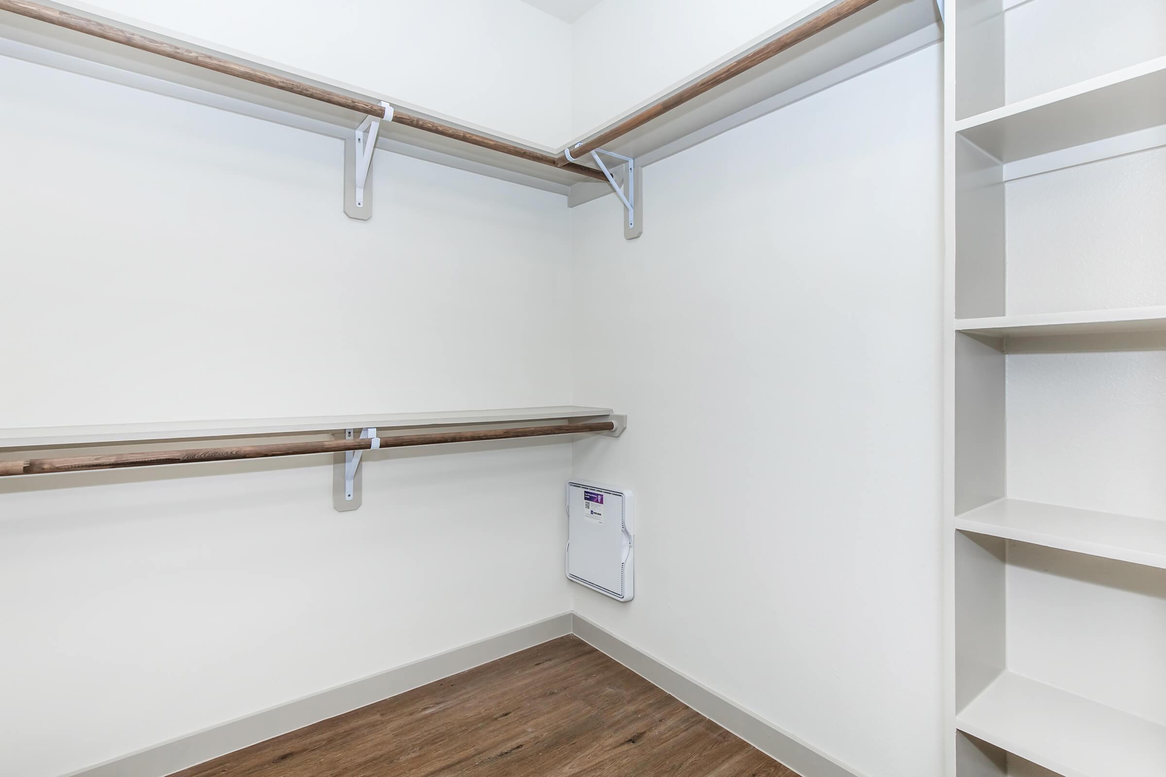 Empty closet featuring two wooden shelves mounted on white walls, with a small white cabinet on the floor and an additional shelving unit on the right side. The flooring is a warm wood finish, contributing to a clean and minimalist aesthetic.