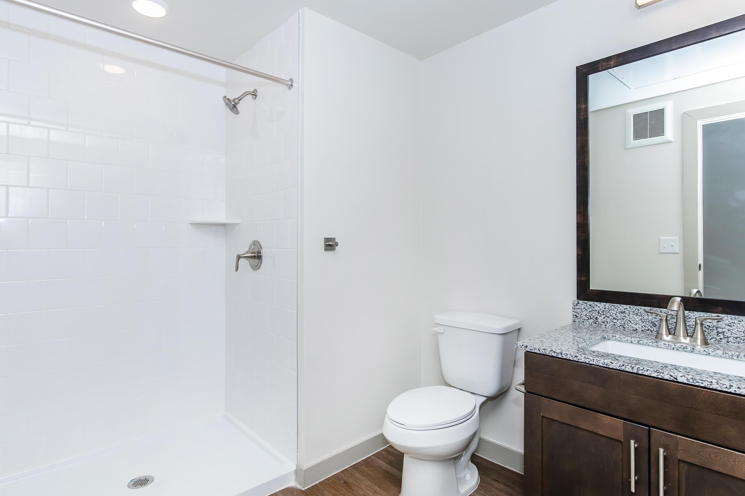 A modern bathroom featuring a white tiled shower with a chrome showerhead, a toilet, and a dark wood vanity with a granite countertop and a round mirror. The walls are painted in a light color, and there is a ceiling light providing bright illumination.