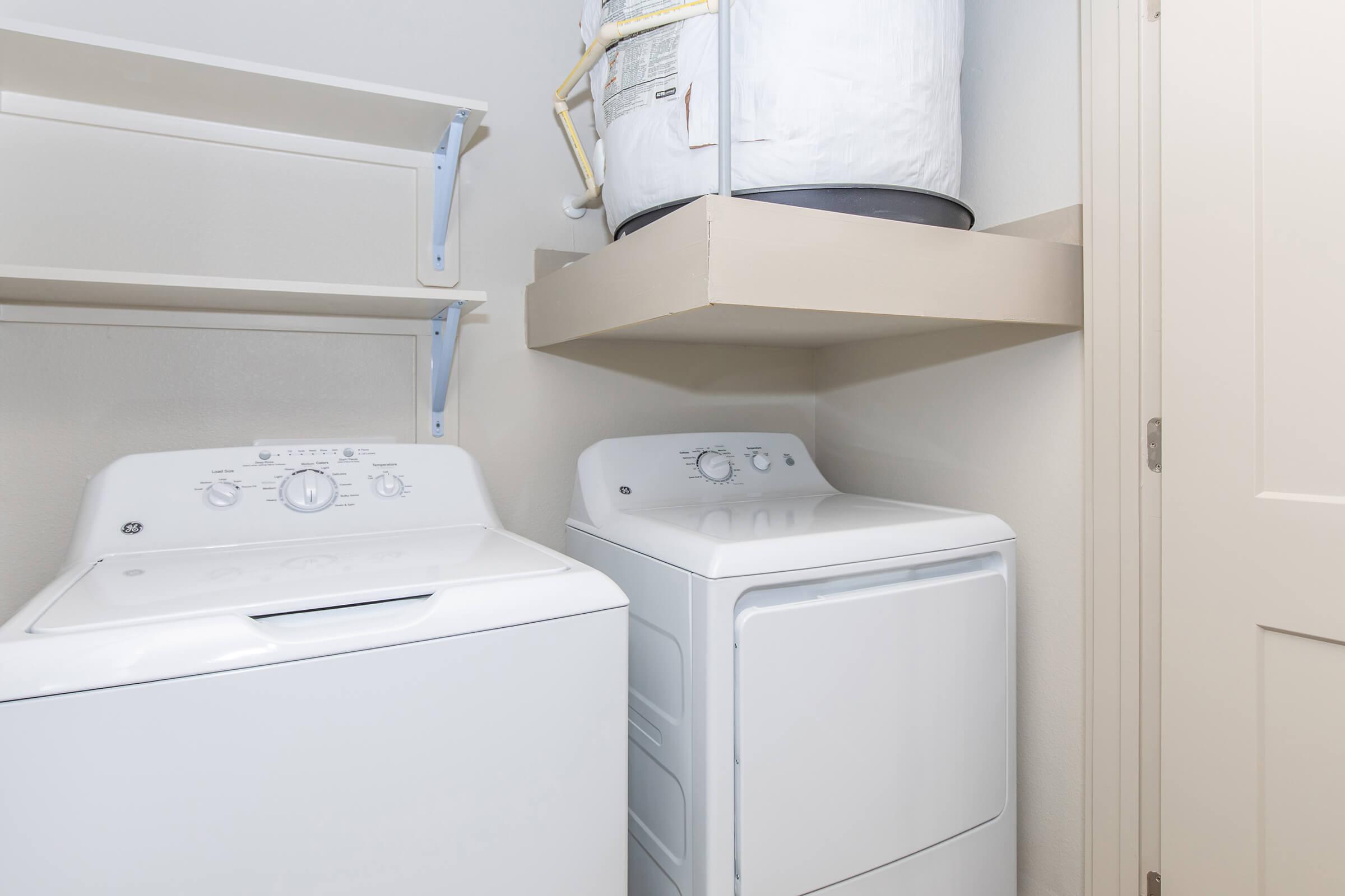 A white washing machine and a white dryer are positioned side by side in a laundry room. Above them, there are two shelves, and a water heater is visible on a shelf mounted against the wall. The room features neutral-colored walls and a closed door.