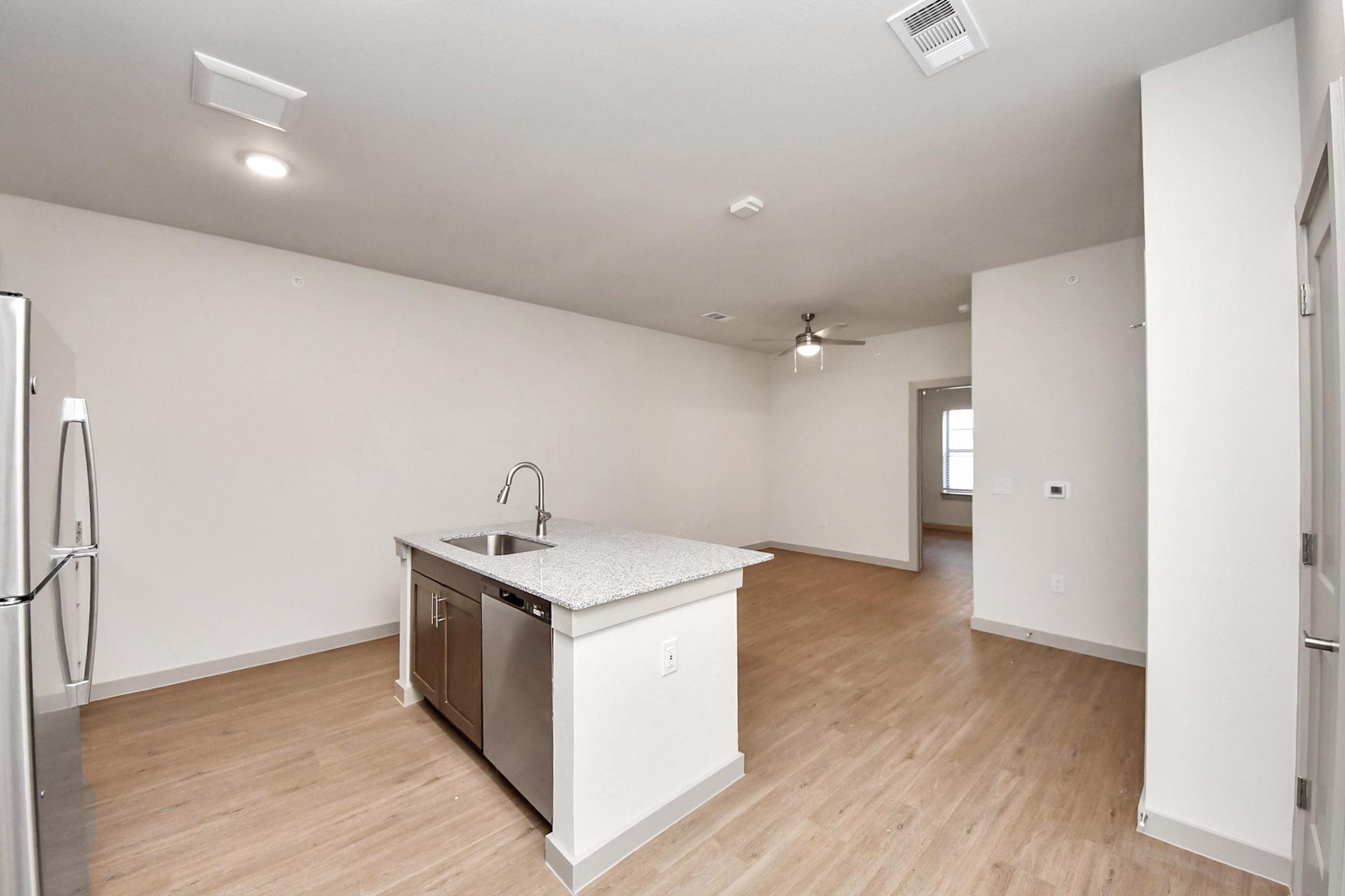 A modern, open-concept living space featuring a kitchen island with a sink, stainless steel appliances, and light wood flooring. The area is well-lit with overhead lights and a ceiling fan. There are minimal furnishings, creating a spacious feel, and windows along one wall allow natural light.