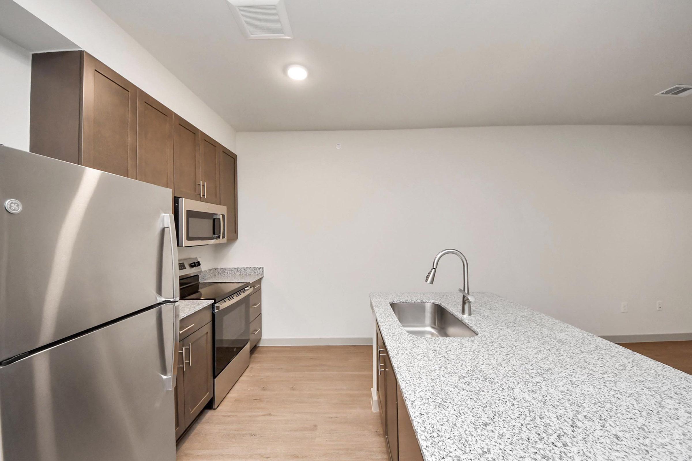 A modern kitchen featuring stainless steel appliances, including a refrigerator, oven, and microwave. The kitchen has wooden cabinetry, a spacious granite countertop with a sink, and a minimalist design. The walls are light-colored, providing a bright and open atmosphere.
