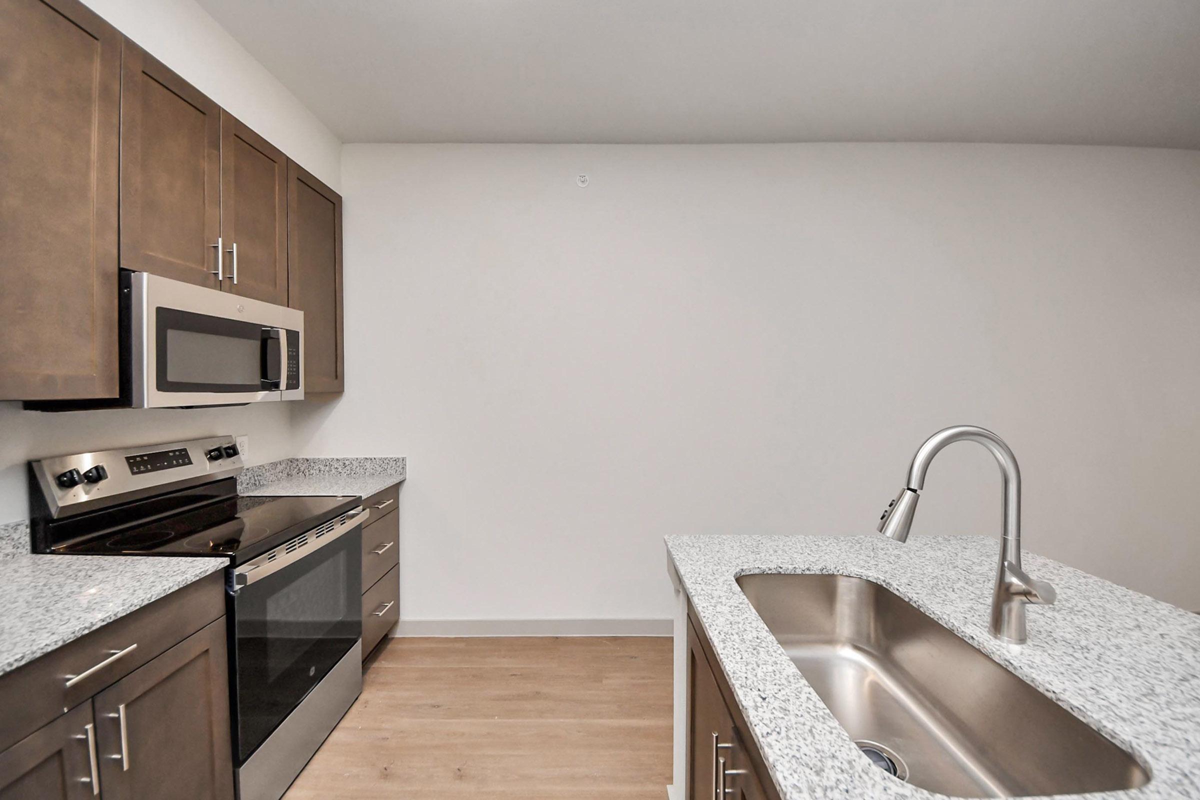 Modern kitchen featuring stainless steel appliances, including an oven, microwave, and sink. The countertops are made of light-colored granite, and the cabinetry is dark wood. The kitchen has a spacious, open design with a clear view of the floor, which is light-colored wood.