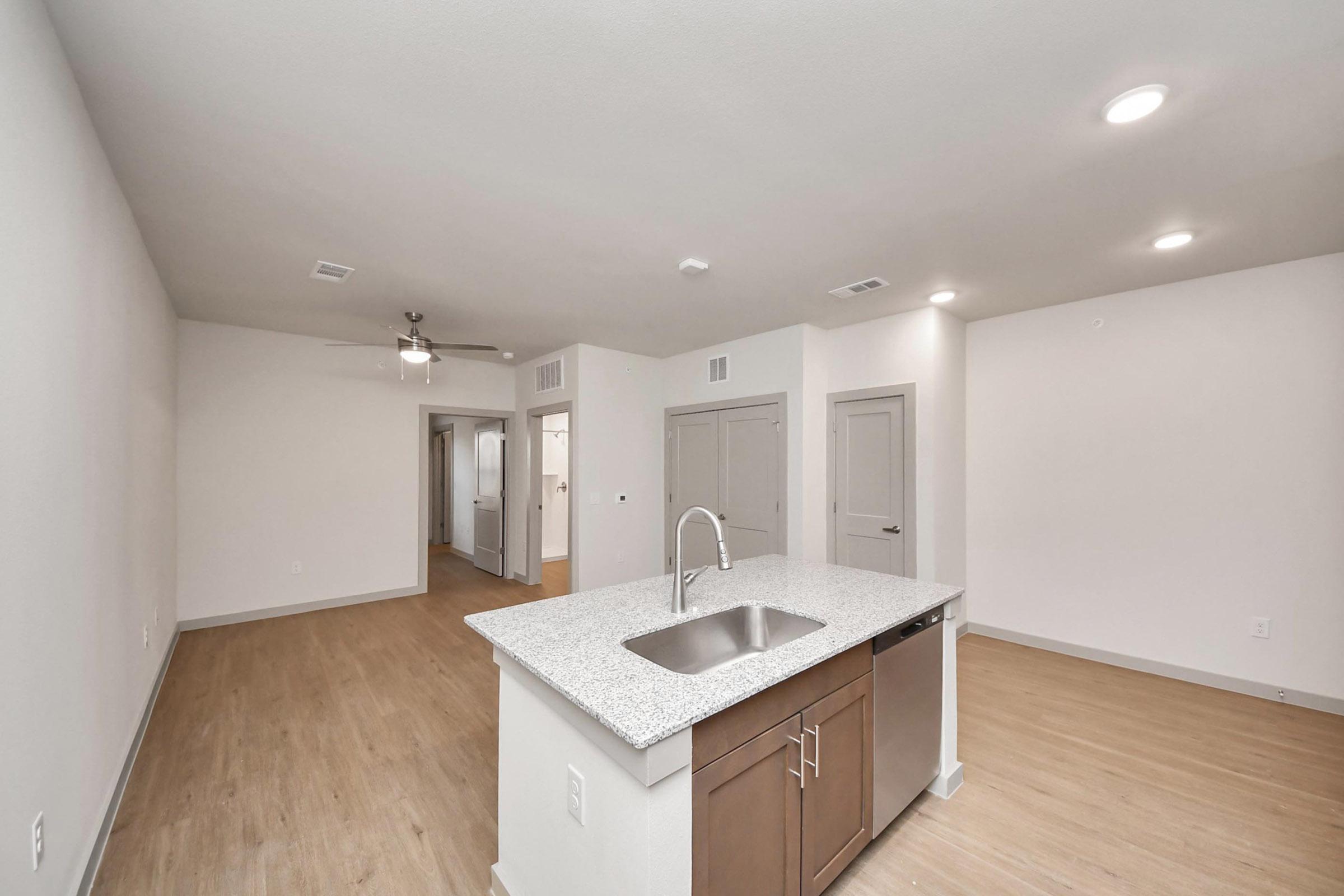 A modern, open-concept living space featuring a kitchen with a granite countertop island, stainless steel sink, and dark wood cabinetry. The area has light-colored walls and wooden flooring, with a ceiling fan and doorways leading to other rooms visible in the background.
