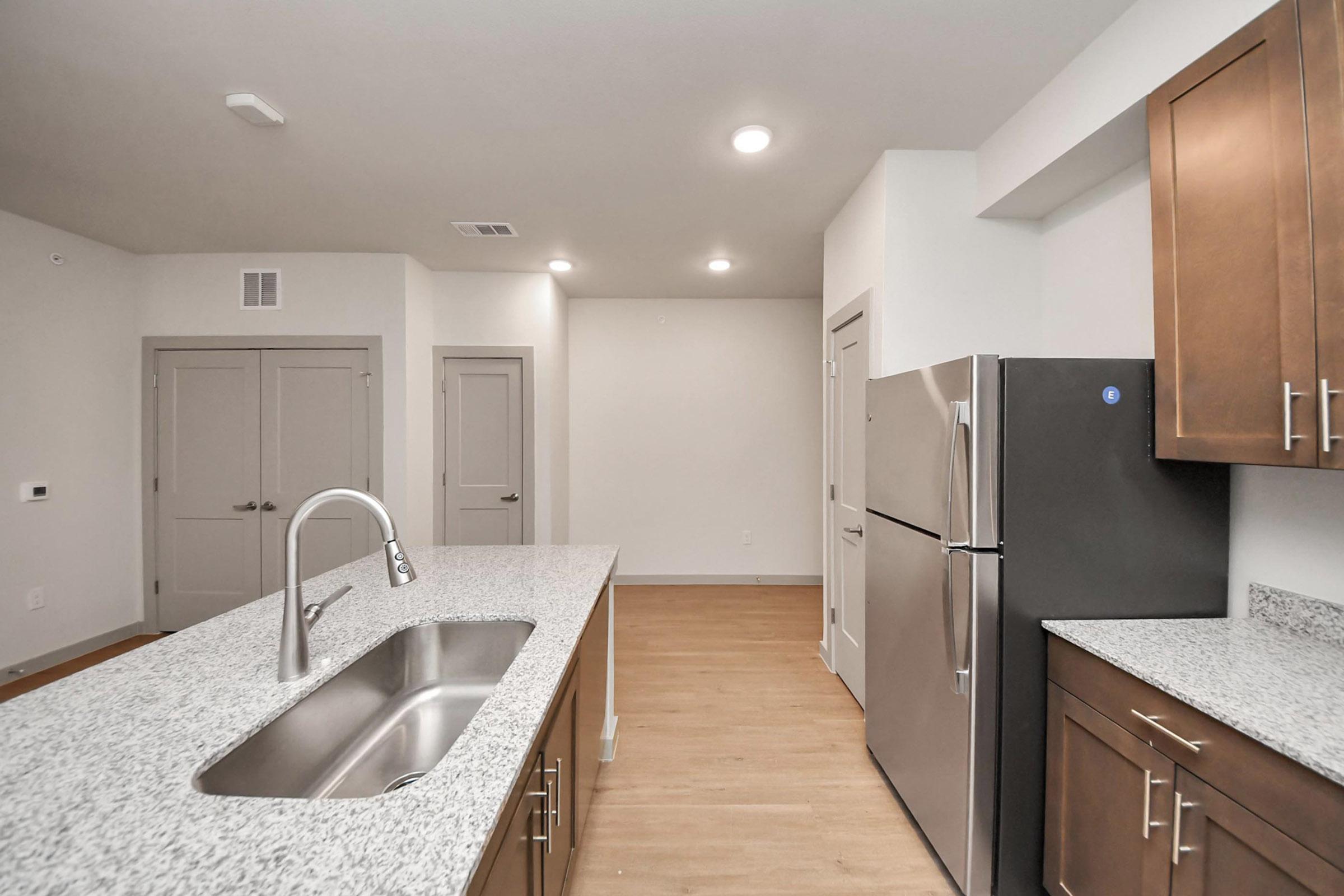 Bright modern kitchen featuring a spacious granite countertop island, stainless steel sink, and sleek refrigerator. The background shows open living space with light-colored walls and doors leading to other rooms, creating a welcoming atmosphere.