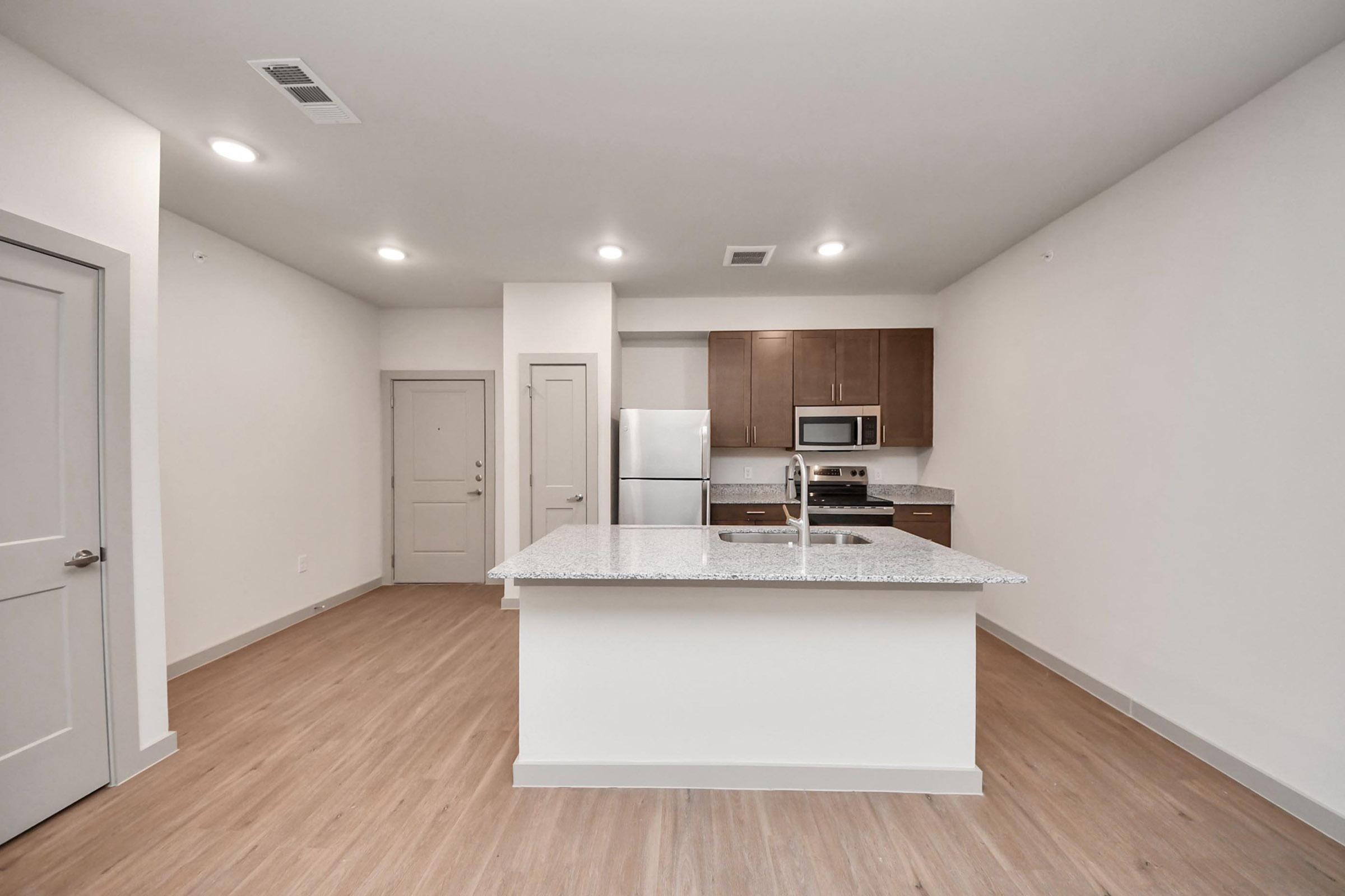 Modern kitchen area featuring a central island with a white granite countertop, stainless steel appliances including a refrigerator and microwave, and wooden cabinetry. The space has light-colored walls and warm wood flooring, with bright overhead lighting and a front door visible in the background.