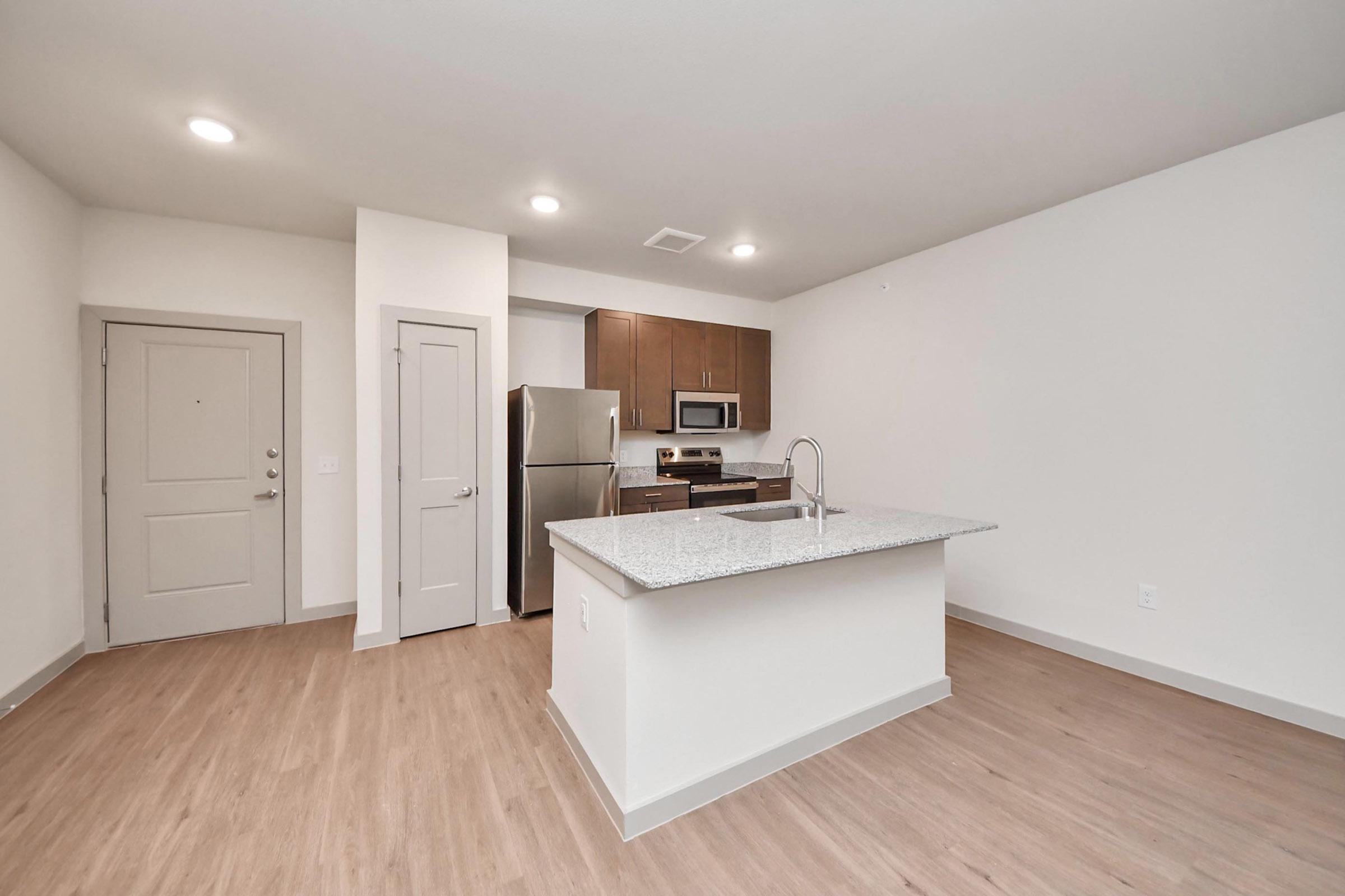 A modern kitchen and living area with light wood flooring. The kitchen features an island with a granite countertop, stainless steel appliances, and wooden cabinets. There is a front door visible, and the space is well-lit with recessed lighting and an open layout.