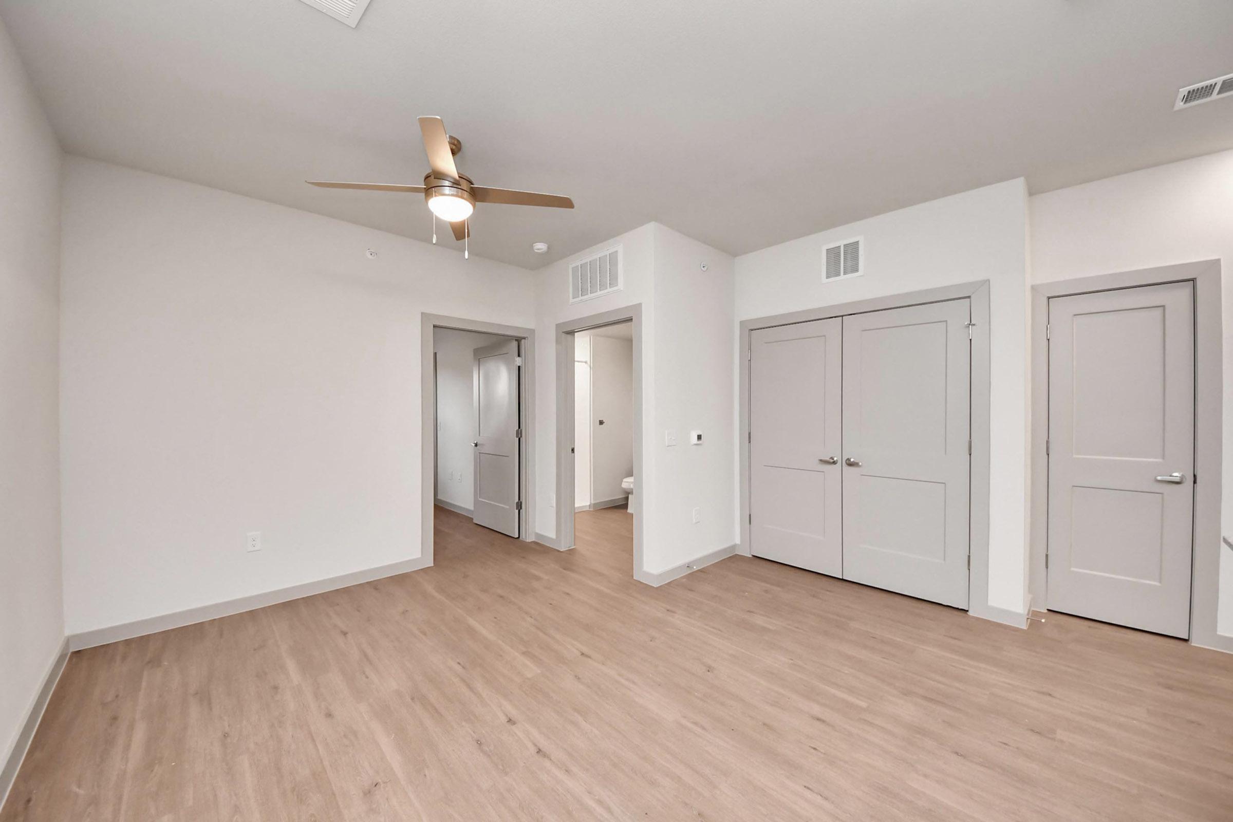 A spacious and empty room featuring light wood flooring, a ceiling fan, and neutral-colored walls. There are two closed doors leading to separate areas, one possibly a bathroom. The overall atmosphere is bright and minimalistic, suitable for various interior design styles.