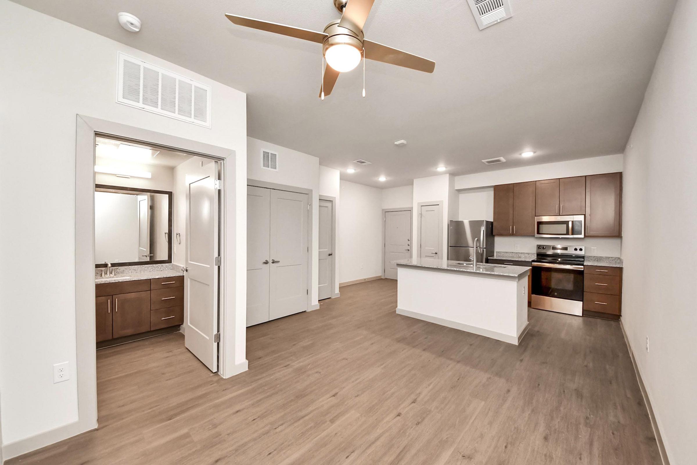 Modern open-concept kitchen and living area featuring wood-style flooring, stainless steel appliances, and warm-toned cabinetry. A ceiling fan hangs above, and a doorway leads to a bathroom with dual sinks and a large mirror. The overall design is sleek and contemporary, with ample natural light.
