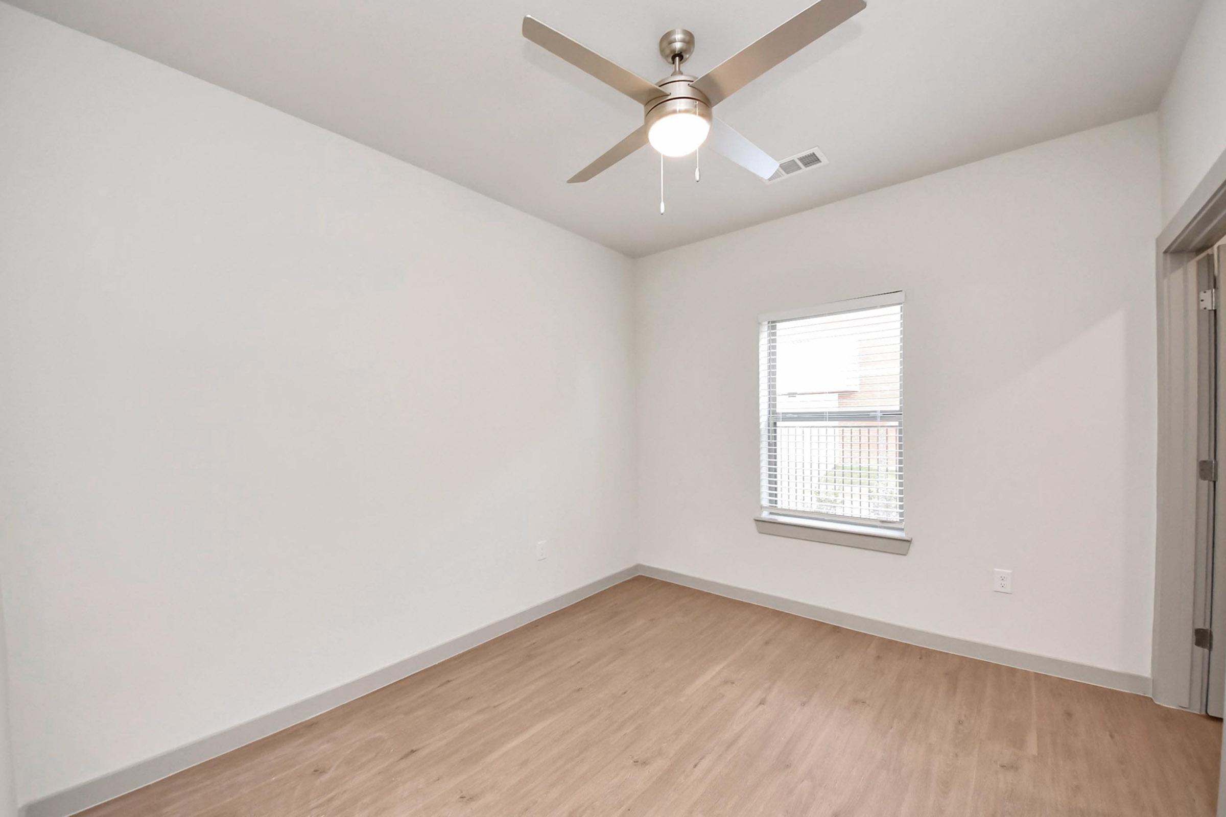 A bright, empty room featuring light-colored walls and wood-like flooring. There is a ceiling fan with a light fixture and a window with blinds, allowing natural light to enter. The overall appearance is modern and minimalistic.