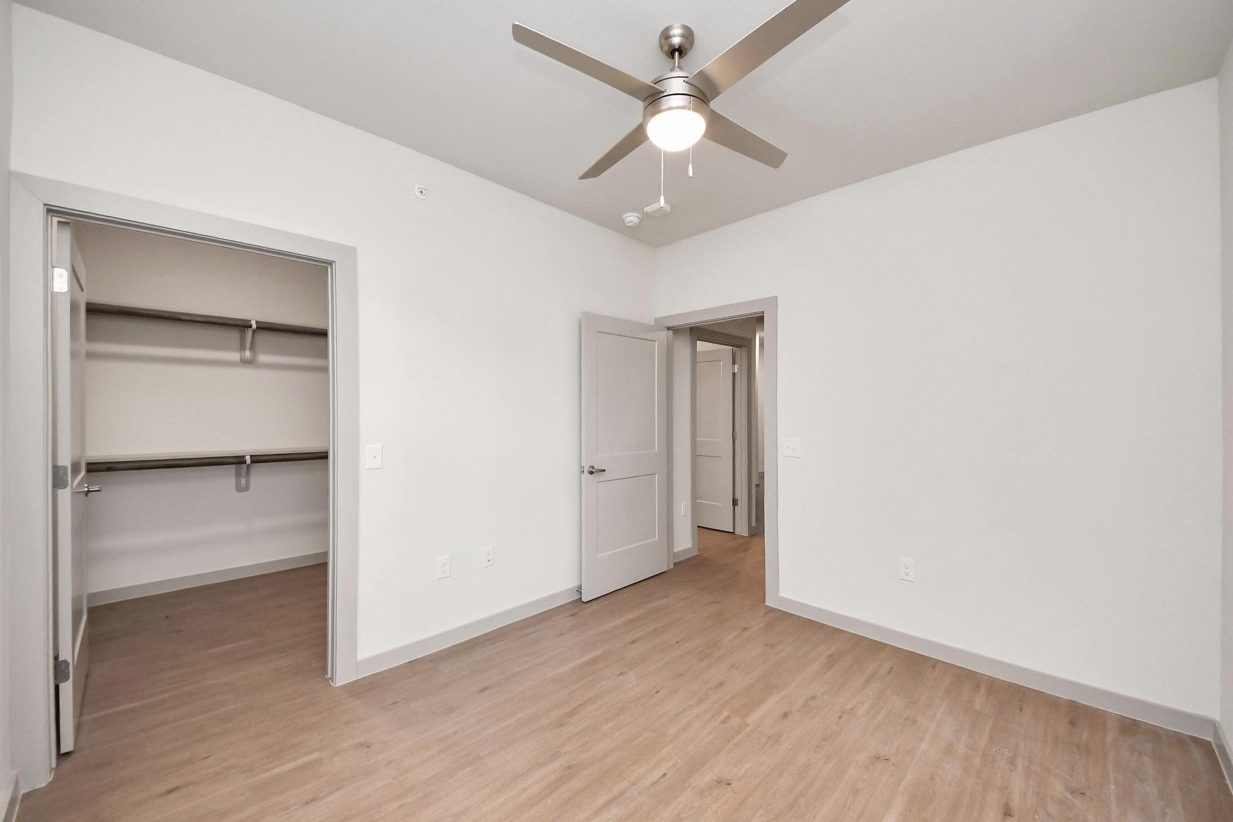 Empty room with light-colored walls and wooden flooring, featuring a modern ceiling fan. A doorway leads to a closet space on one side, while another door provides access to an adjacent room. Soft, natural light fills the space, creating a bright and airy atmosphere.