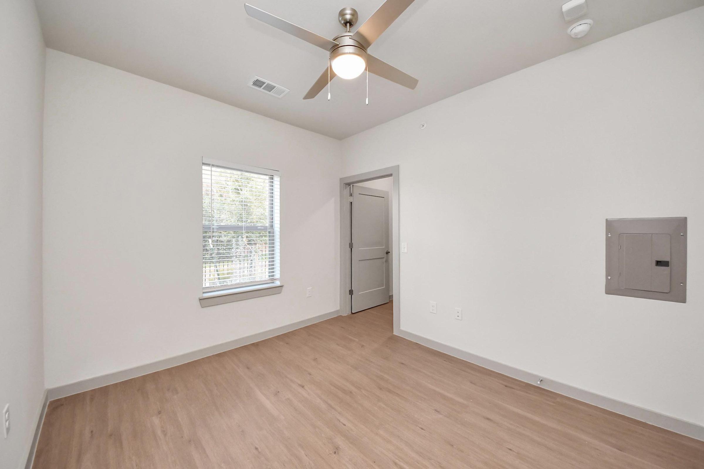 Empty room with light-colored walls and wooden flooring. A ceiling fan is mounted above, and there is a window with blinds allowing natural light in. A door on the right leads to another space, and a utility panel is mounted on the wall, indicating a modern interior design.