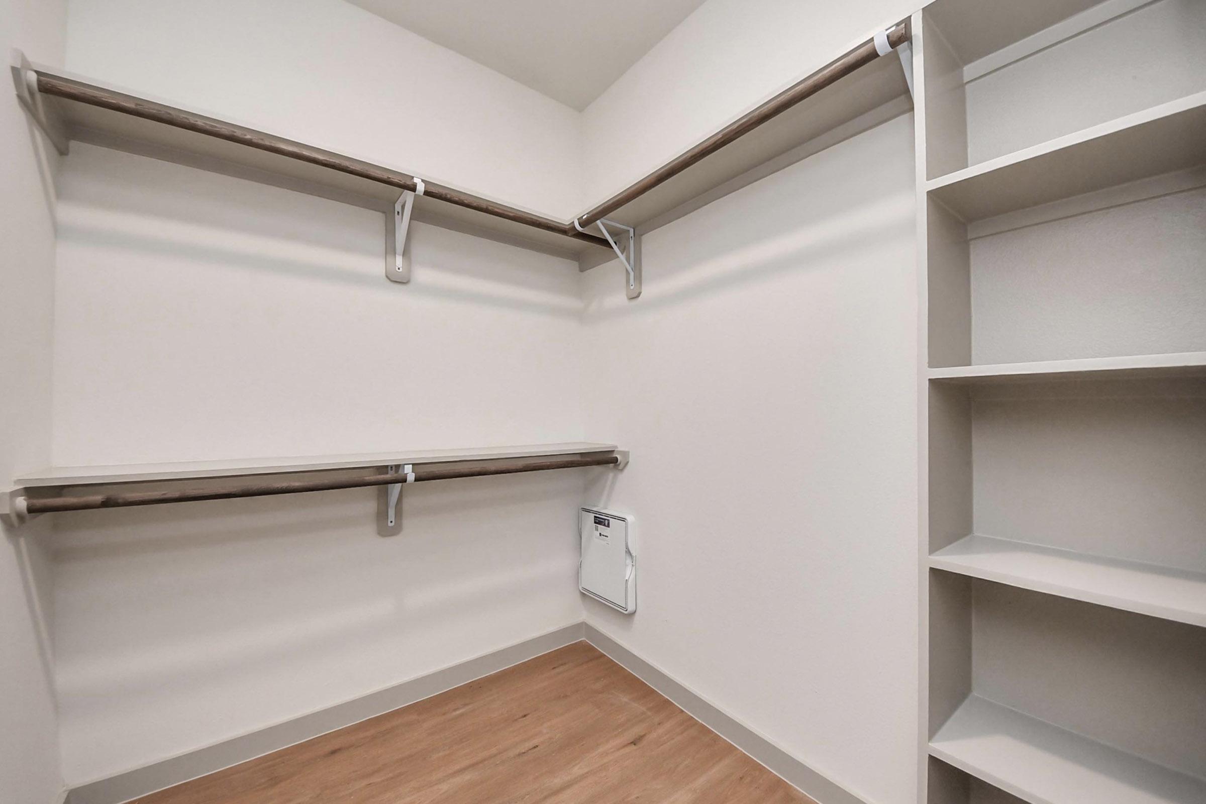 Empty closet with two horizontal shelves mounted on the walls, light-colored walls, and a wooden floor. There is a small white access panel on the lower wall, and a tall shelf unit on the right side. The overall space is uncluttered and bright, suitable for storage or organization.