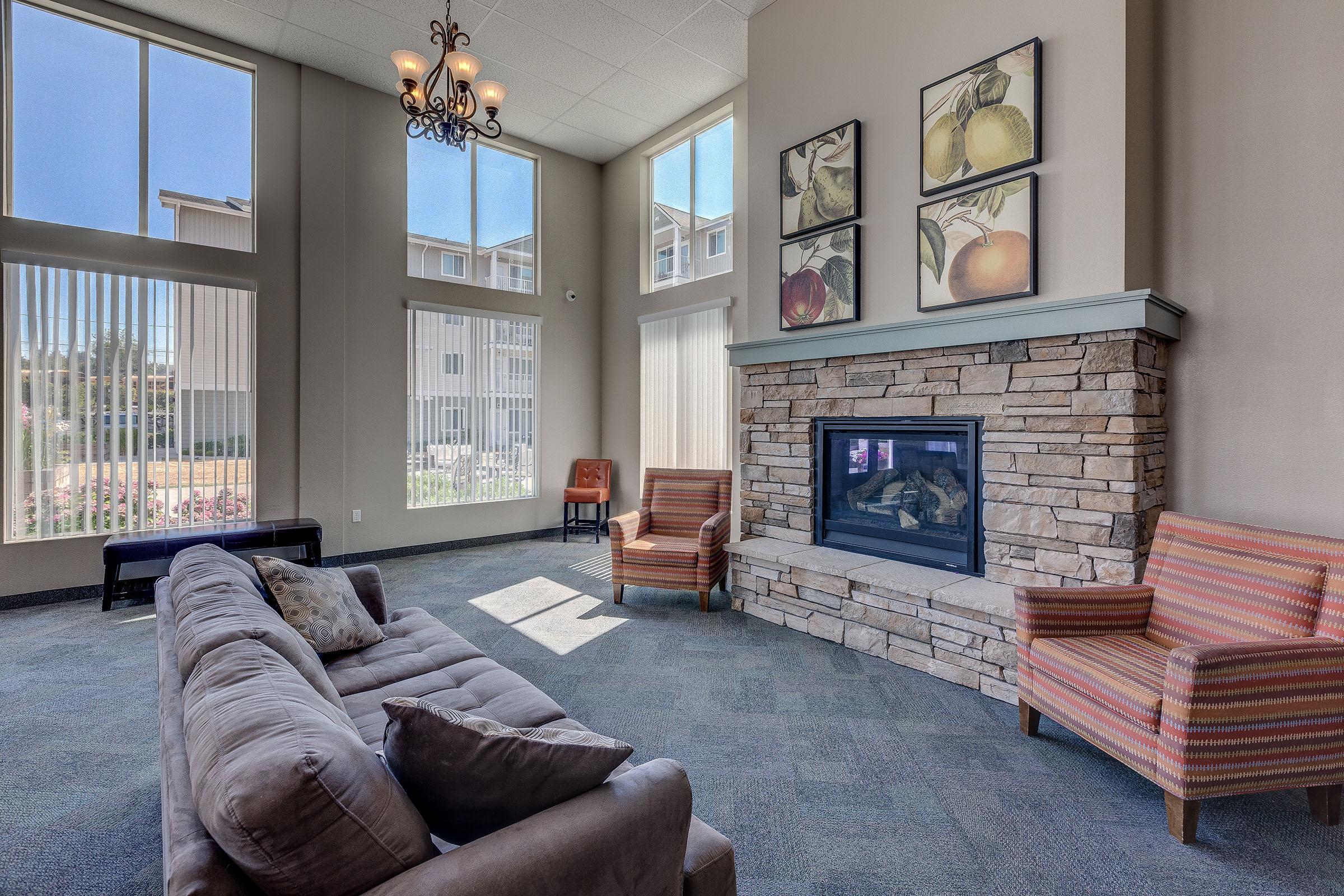 A cozy living room with a stone fireplace as the focal point. It features a gray sofa, two patterned armchairs, and large windows allowing natural light. Artwork of fruit hangs on the walls, and the floor is carpeted, creating a warm and inviting atmosphere.