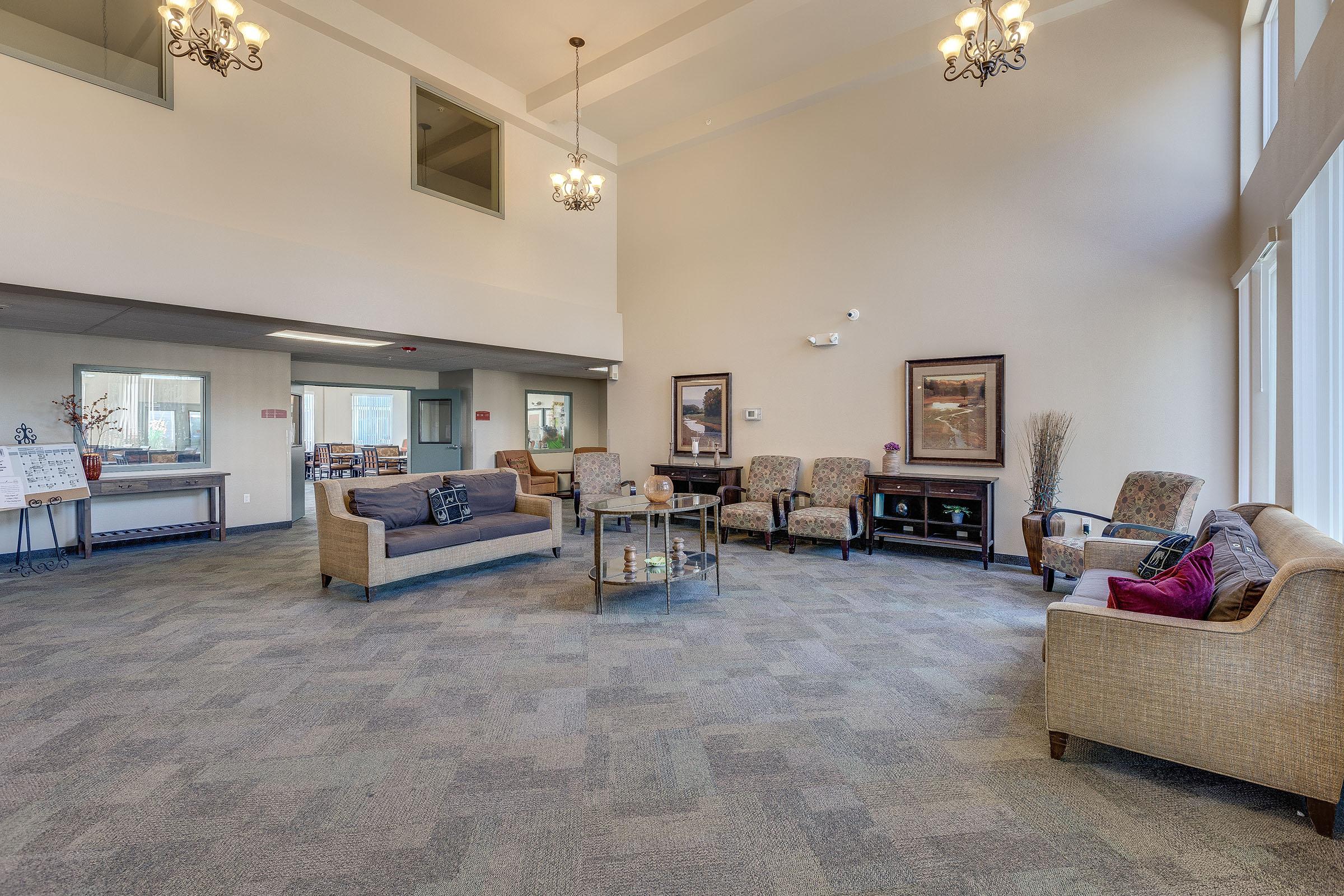 A spacious, bright lounge area featuring comfortable seating arrangements, including sofas and armchairs. Large windows allow natural light to fill the space, adorned with decorative art pieces. The carpeted floor complements the inviting atmosphere, creating a welcoming environment for relaxation or social gatherings.