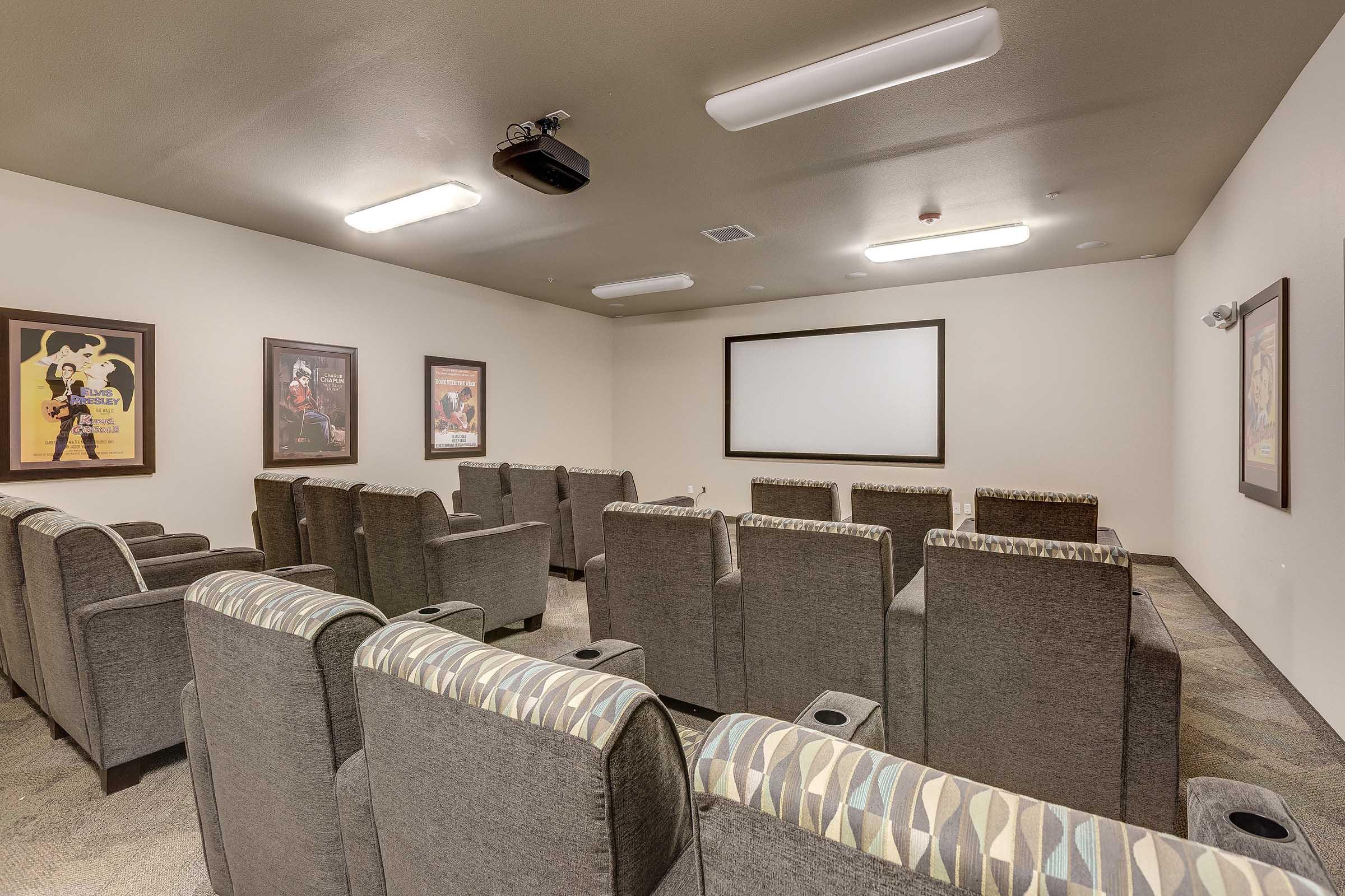 A modern home theater setup featuring rows of plush, gray recliner chairs with armrests, equipped with cup holders. The walls are adorned with movie posters, and there is a large projection screen at the front. The room is well-lit with overhead lights, creating a cozy movie-watching atmosphere.