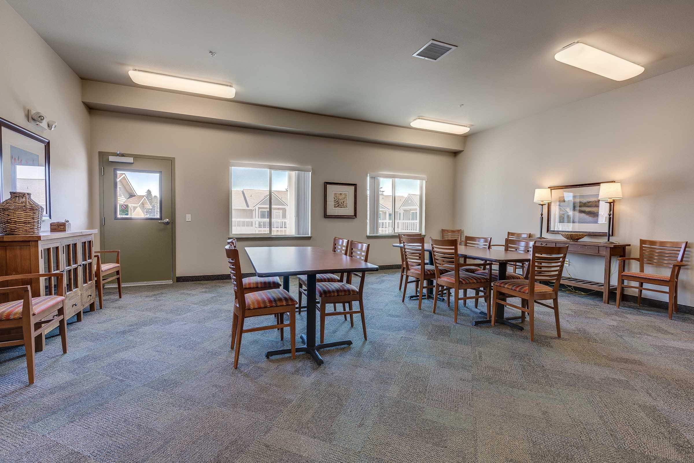 A spacious common area featuring a wooden table surrounded by chairs, a side cabinet, and two large windows allowing natural light. The walls are painted in a light color, contributing to a bright and inviting atmosphere.