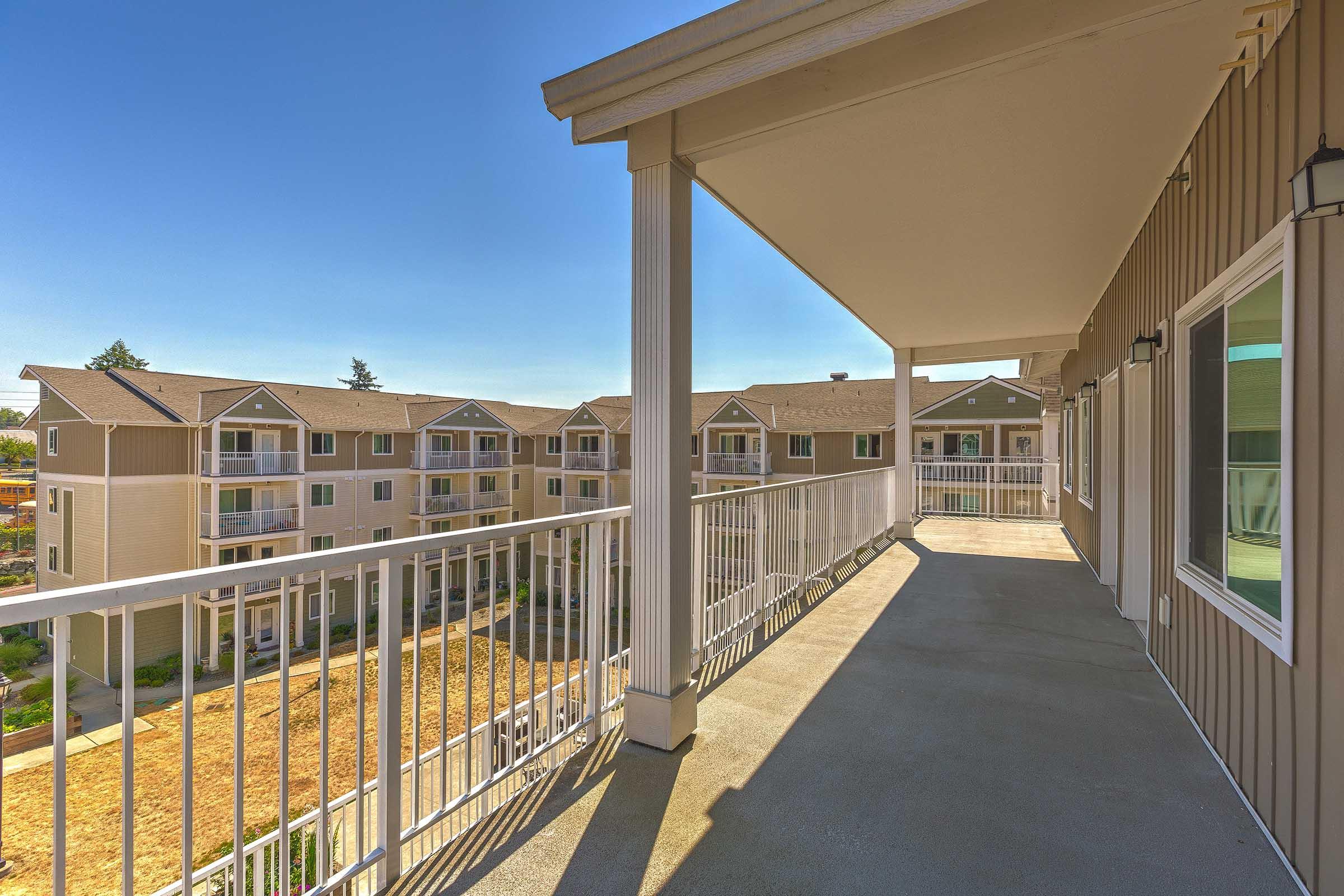 View from a balcony overlooking a courtyard with several multi-story apartment buildings. The bright blue sky and sunlight create a warm atmosphere, highlighting the architectural details of the buildings and the railing of the balcony. The setting conveys a sense of community and open space.