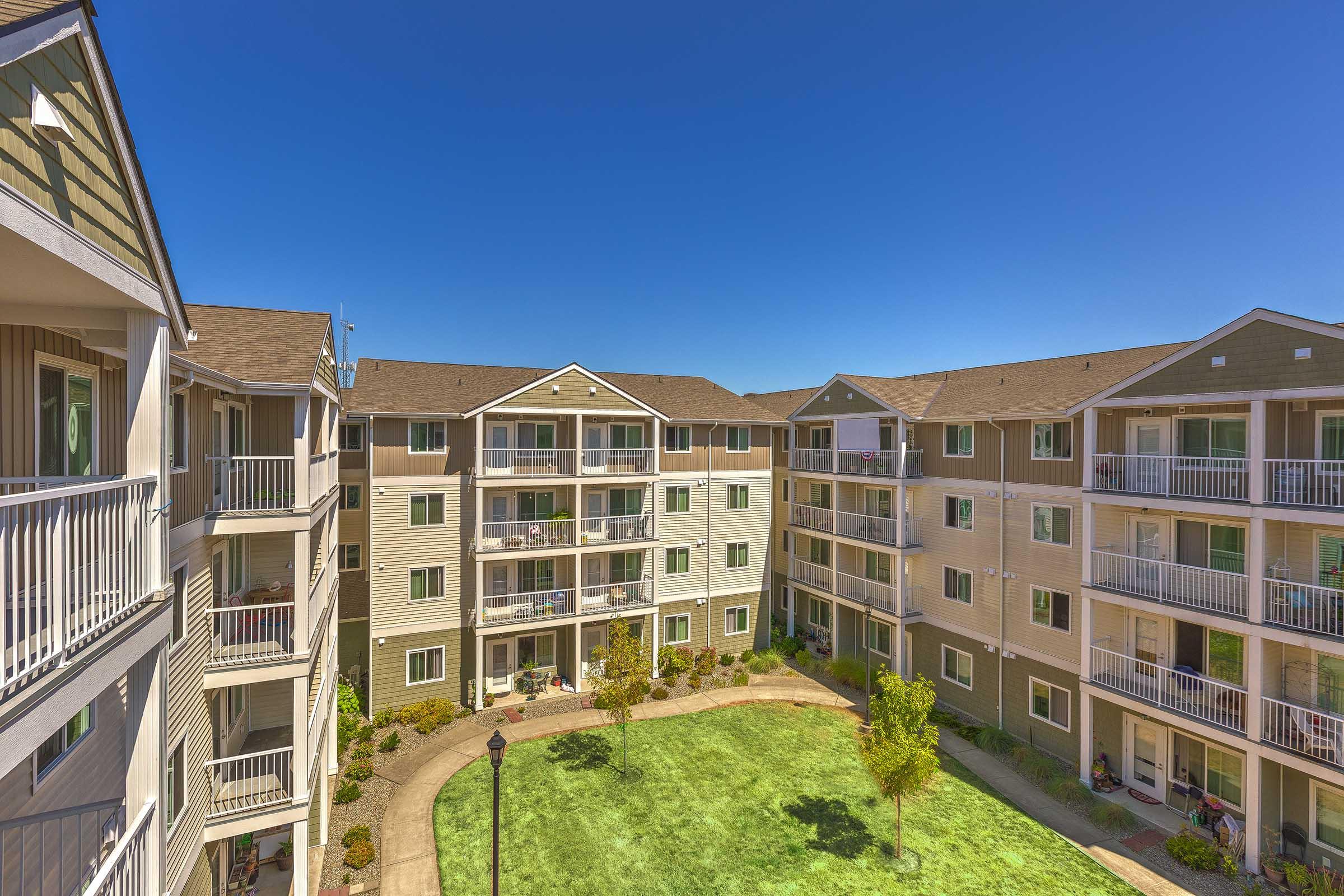 A courtyard view of a multi-story apartment complex with multiple buildings surrounding a green lawn. The buildings feature balconies, and residents can be seen on some of the patios. The sky is clear and blue, creating a bright and welcoming atmosphere.
