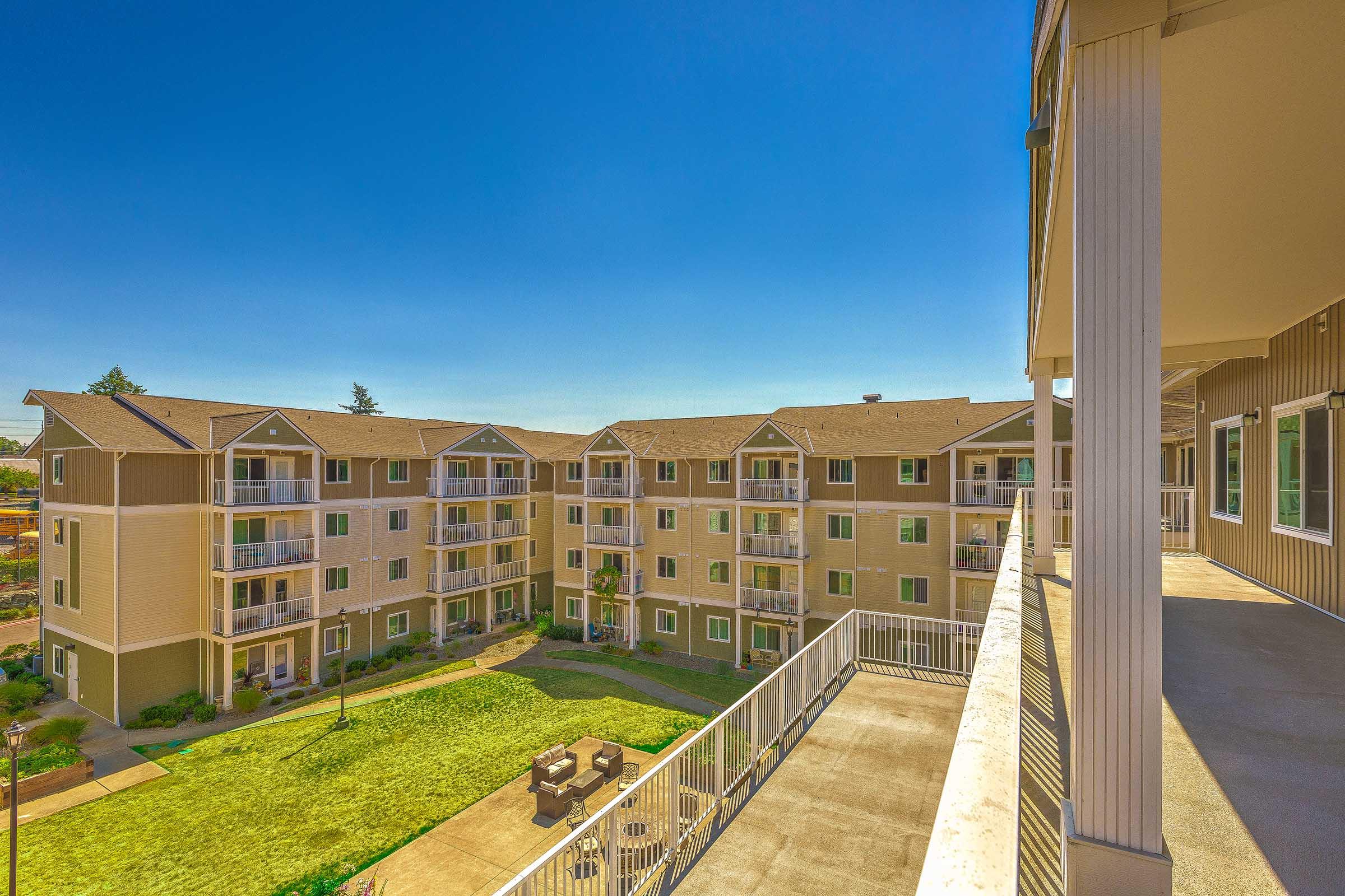 A view of a multi-story apartment complex with multiple balconies, surrounded by green grass and landscaping. The sky is clear blue, and the image captures the building's modern architecture and outdoor communal spaces.