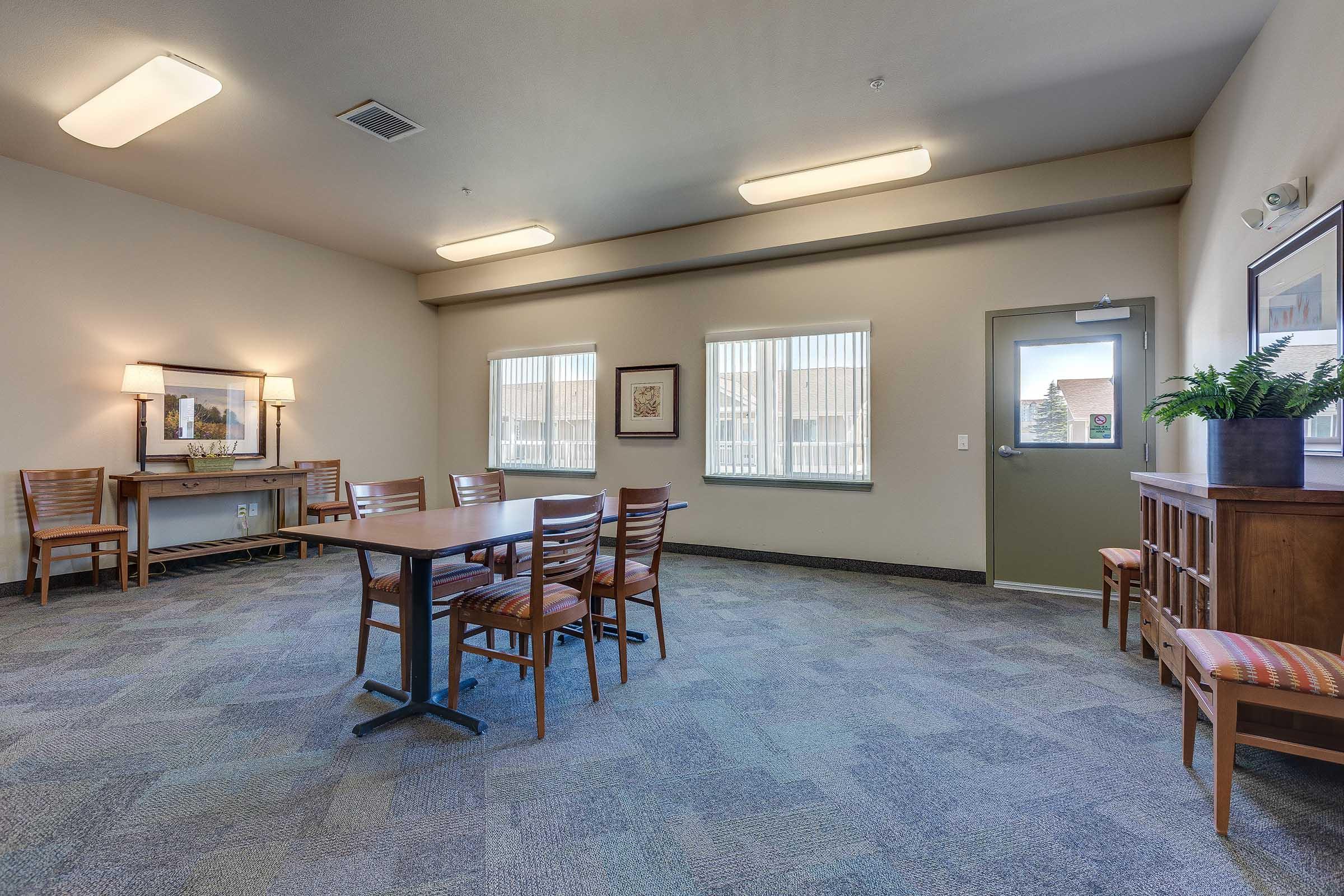 Interior of a communal room featuring a large table surrounded by chairs, with windows allowing natural light. The walls are painted in neutral tones, and there is a plant on a side table. Lighting fixtures on the ceiling provide bright illumination.