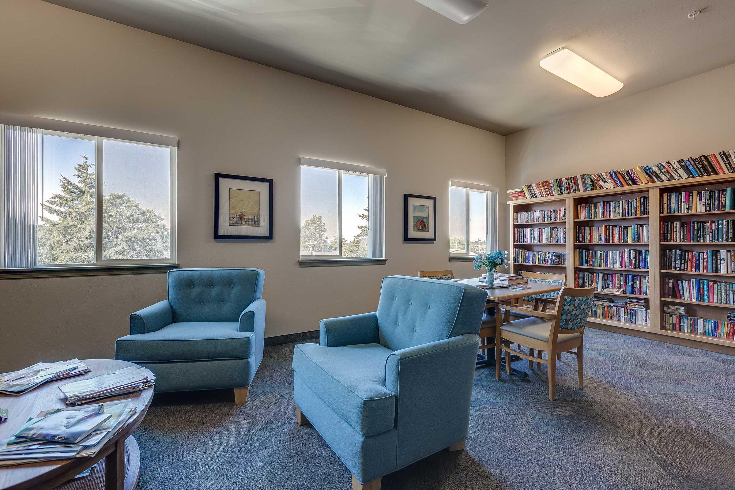 A cozy library space featuring two light blue armchairs, a wooden table, and a bookshelf filled with books. Large windows let in natural light, and there are framed pictures on the walls. Magazines are stacked on a nearby table, contributing to a welcoming reading environment.