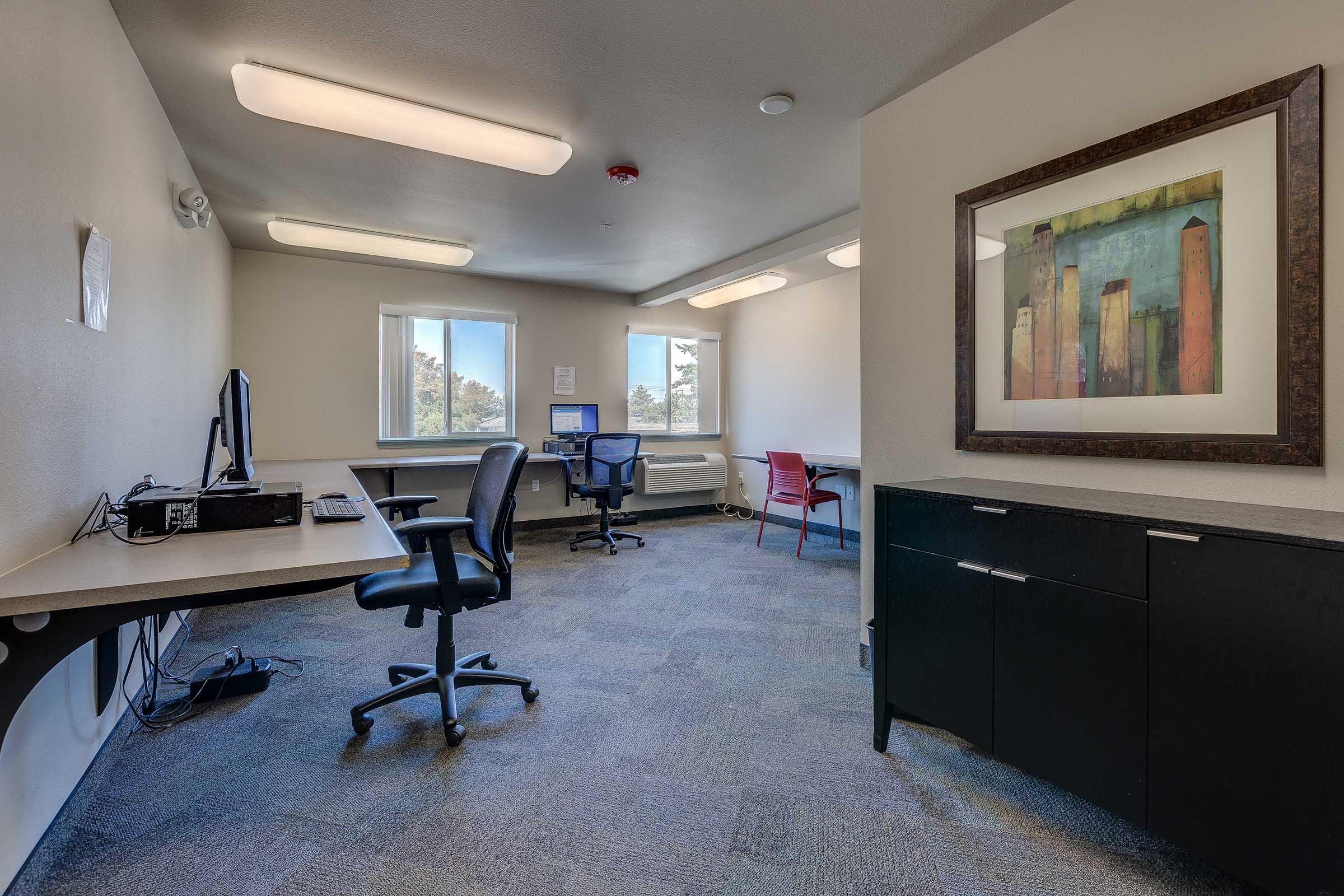 An empty office space featuring two desks with chairs, a computer setup, and a large framed artwork on the wall. It has windows allowing natural light to enter and a carpeted floor. The neutral-colored walls and simple furniture create a minimalist ambiance.