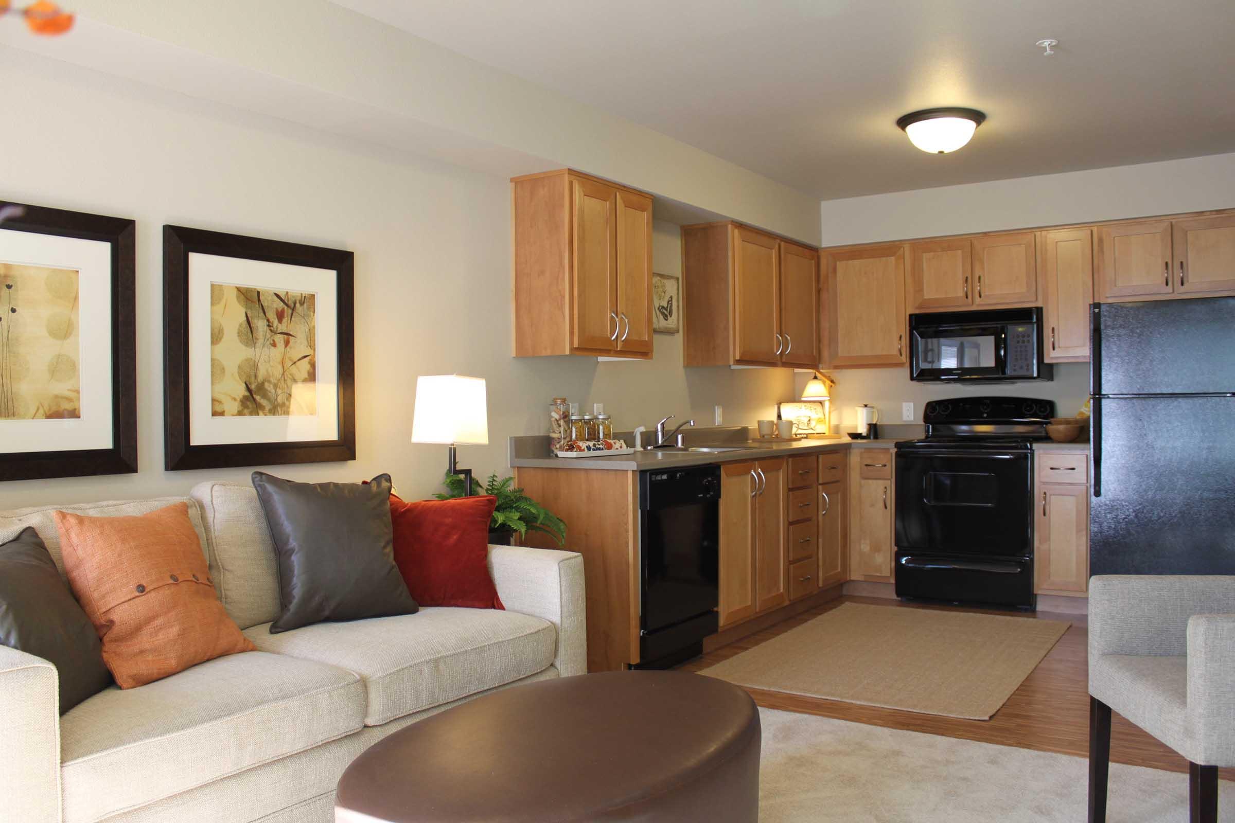 A cozy living area featuring a light-colored sofa with colorful cushions, a small round table, and framed artwork on the walls. Adjacent is a kitchen with wooden cabinets, a black refrigerator, and modern appliances. Soft lighting creates a warm atmosphere, with a rug on the floor.