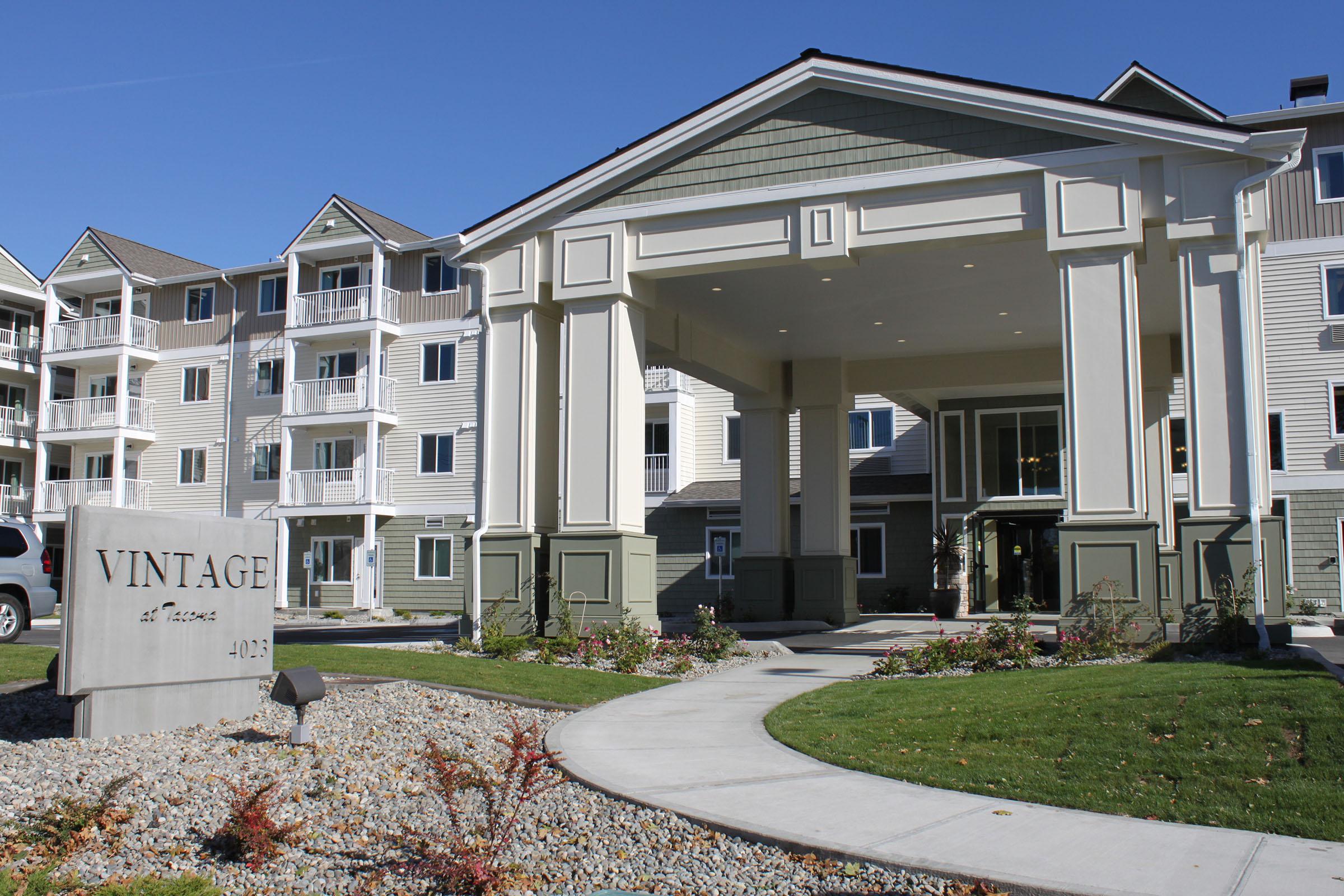 Exterior view of a multi-story residential building with a modern design. The entrance features a large awning and decorative columns. A sign reading "VINTAGE at a glance" is visible near the entrance, surrounded by landscaped greenery and a walkway leading to the door. Clear blue sky in the background.