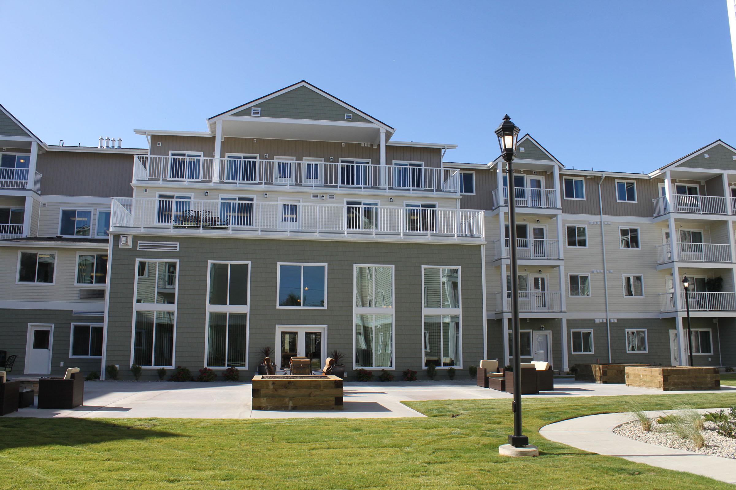 A modern multi-story apartment building with balconies and large windows. The exterior features a light color palette with gray accents. In the foreground, there is a landscaped courtyard with grass and a paved area, including seating arrangements and decorative elements. Clear blue sky overhead.