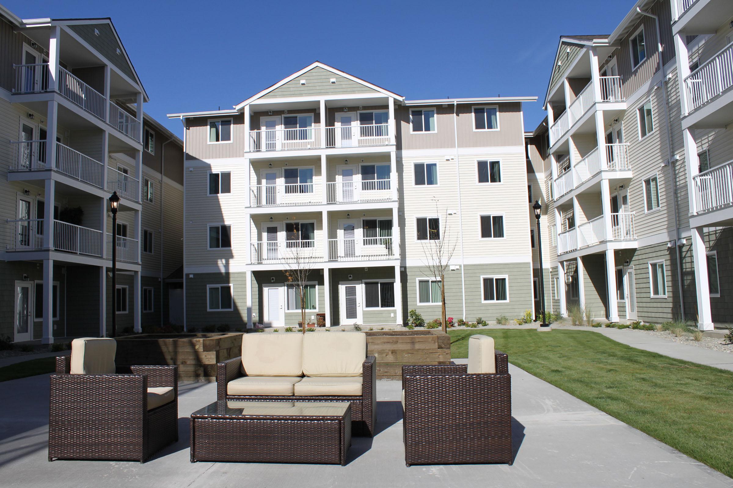 A modern residential complex featuring three multi-story buildings. In the foreground, there is a seating area with a beige couch and dark wicker chairs, surrounded by landscaped grass and trees. The buildings have balconies and a contemporary design, set against a clear blue sky.
